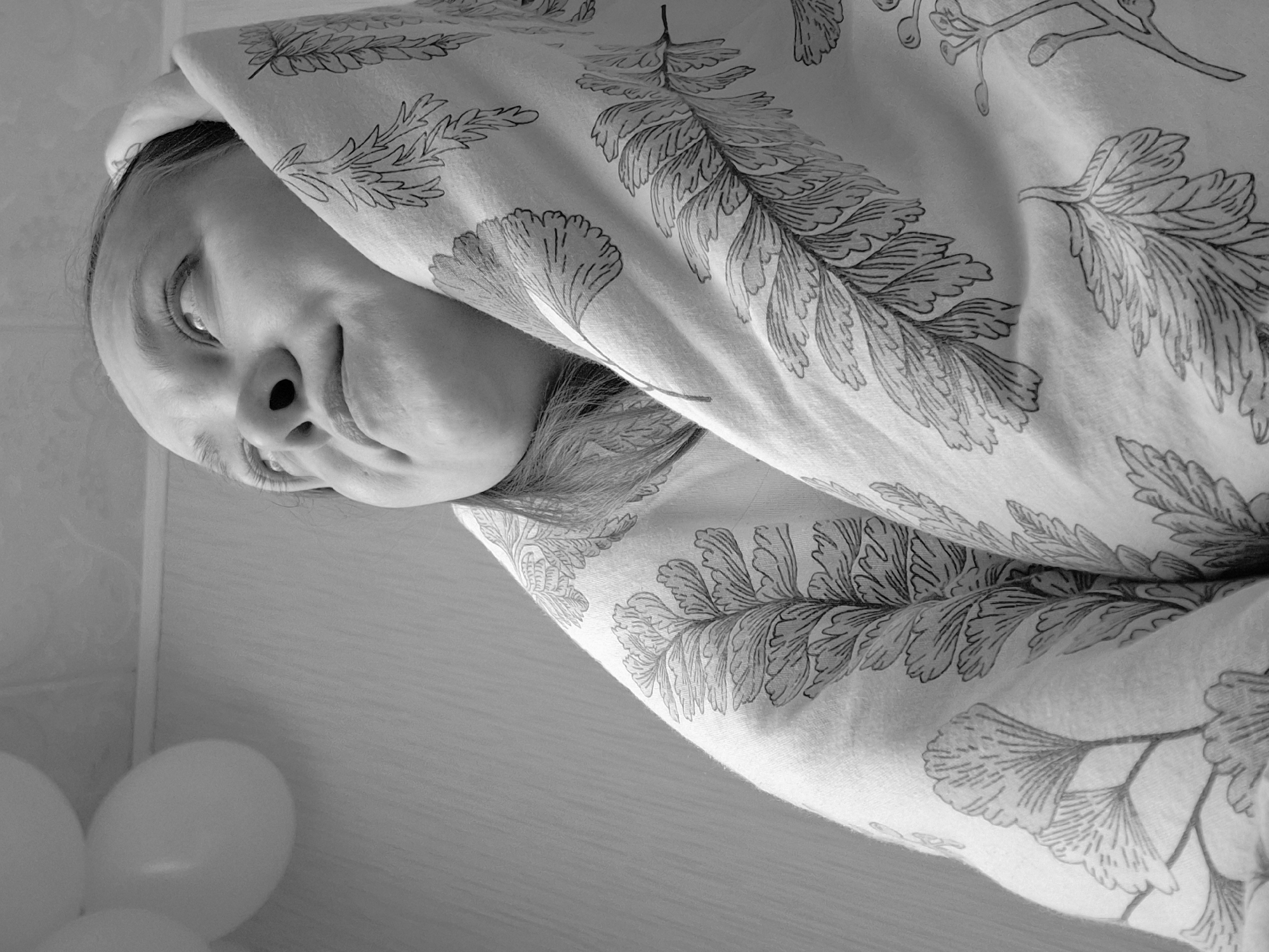 A grayscale, rotated image depicts a person partially concealed under a patterned fabric. The individual, likely a young woman, is looking up and to the left. Only her face and part of her upper body are visible beneath the fabric, which is decorated with botanical motifs. To the lower left of the image, partially visible are several balloons. The location is not discernible from the image itself, although it can be inferred from external information provided that the image may have been taken in the city of Chaikovsky, Russia.