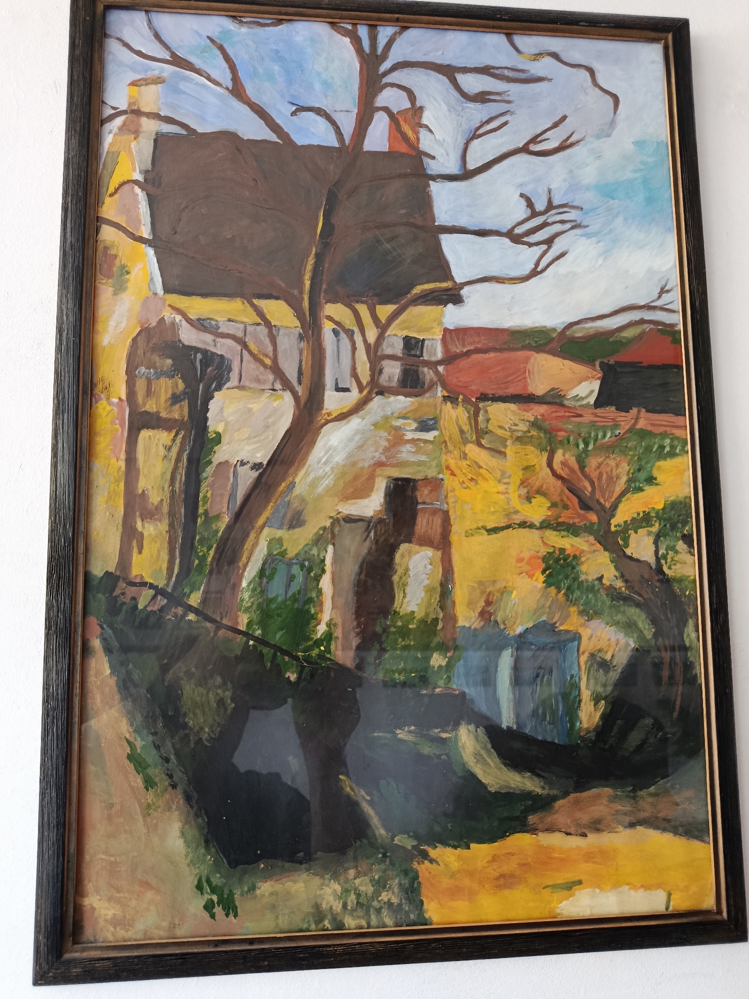 This is a framed oil painting of a house and trees. The style of the painting is impressionistic, with visible brushstrokes and a vibrant color palette. The house is depicted with yellow walls and a brown roof. Bare tree branches extend across the sky, which is a mix of light blue and white. The foreground features dark green foliage and what appears to be a path or walkway. The painting is hanging on a white wall. There are no people in the painting. The painting is not specifically identifiable as being in Braslaw, Belarus.