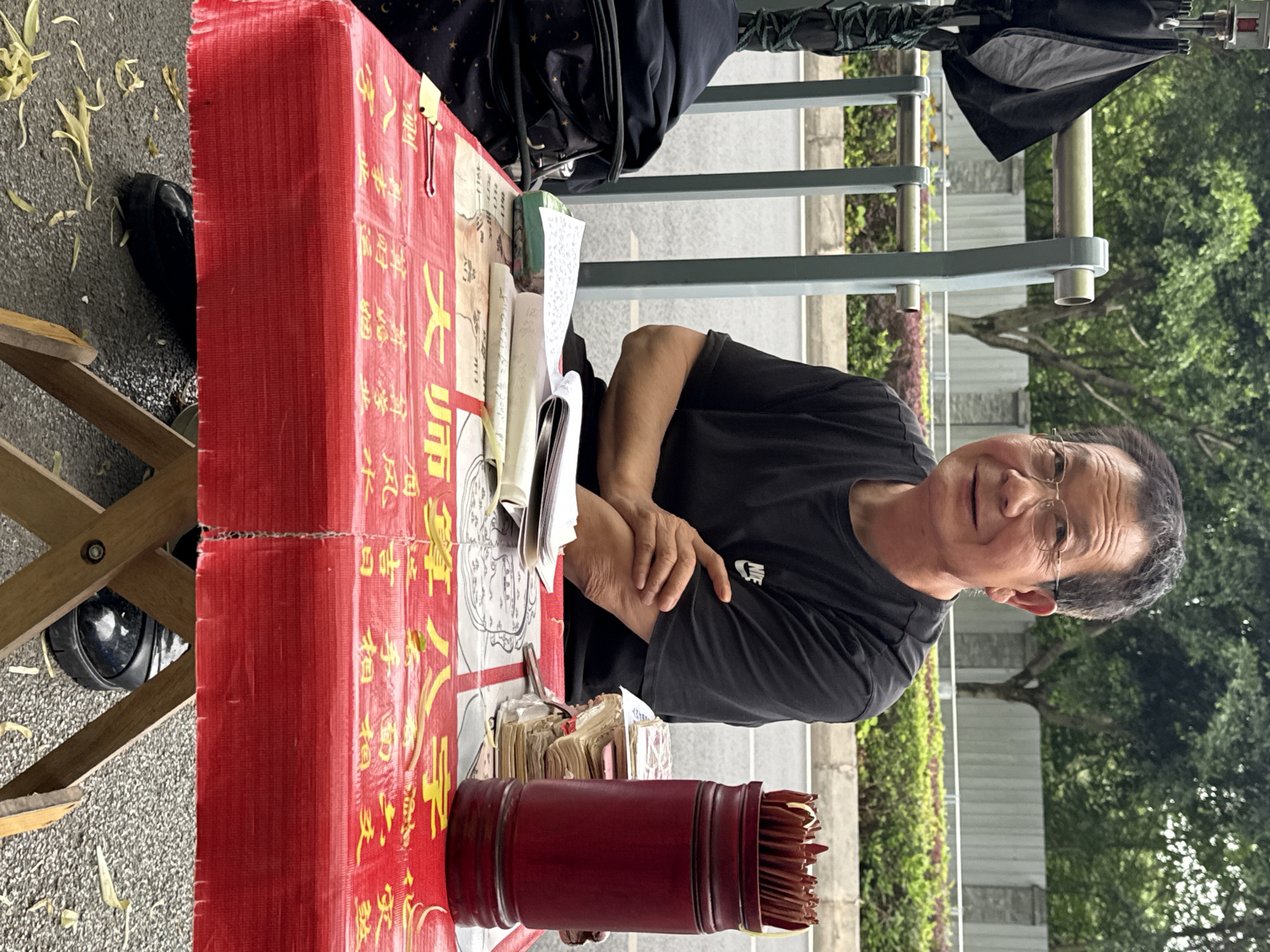 A man sits at a table set up outdoors in what appears to be a public space. The scene is likely in Chongqing High-tech Zone, China. He wears a black t-shirt with a Nike logo and glasses. His arms are crossed, and he looks toward the viewer. The table is covered in a red cloth with yellow Chinese characters. On the table are several books, papers, and a red cylindrical container filled with fortune-telling sticks. A bag hangs on the table behind him, and an umbrella is positioned nearby. In the background, there is a metal railing, greenery, and trees. The ground appears to be paved.