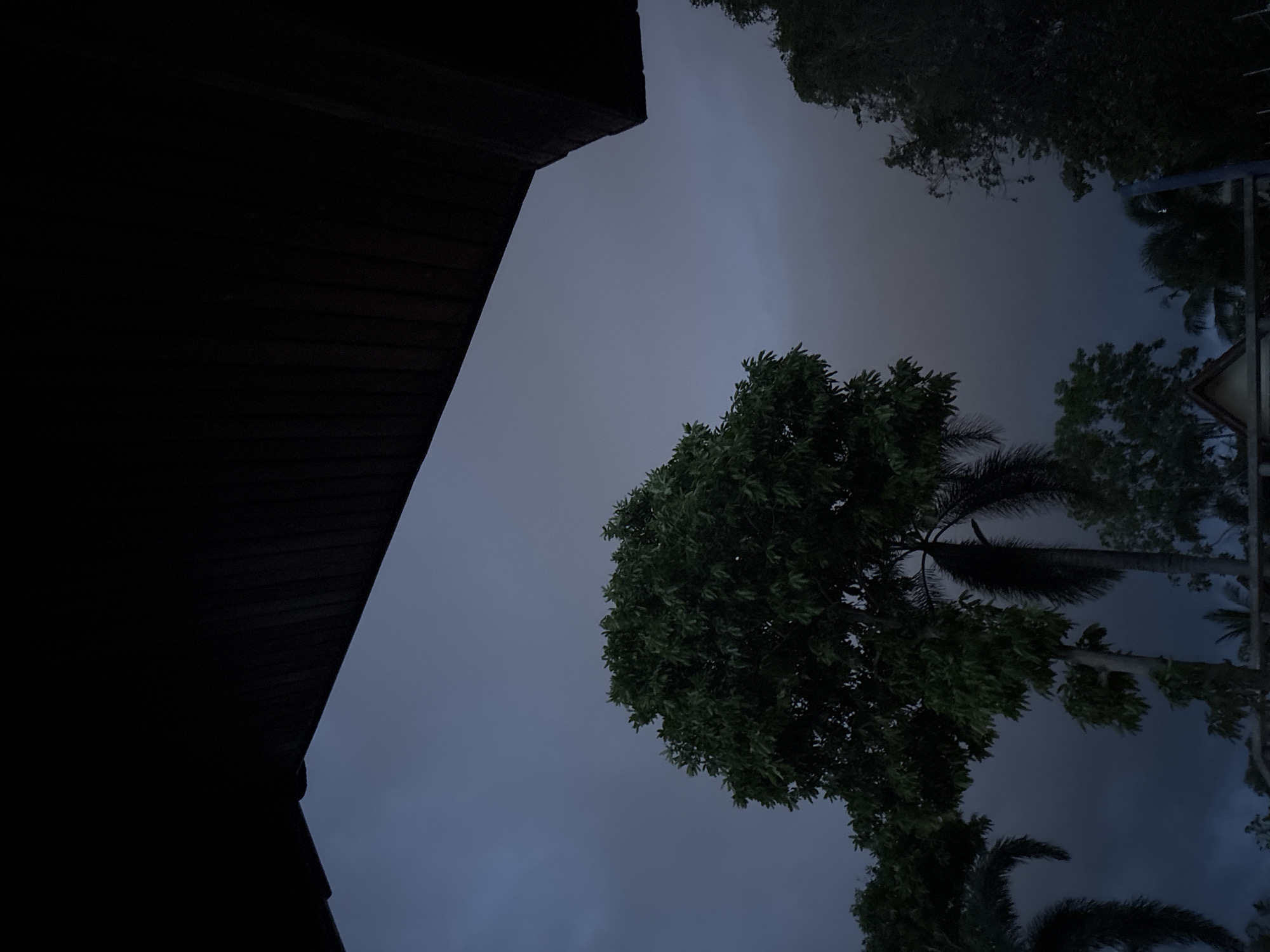 A low-angle shot depicts a dark and cloudy sky in บ้านบางรักษ์, Thailand. A dark-colored structure with a sharp corner occupies the left side of the frame. In the center are trees with dense foliage. Palm trees are visible among the trees. A signpost is barely visible on the right.