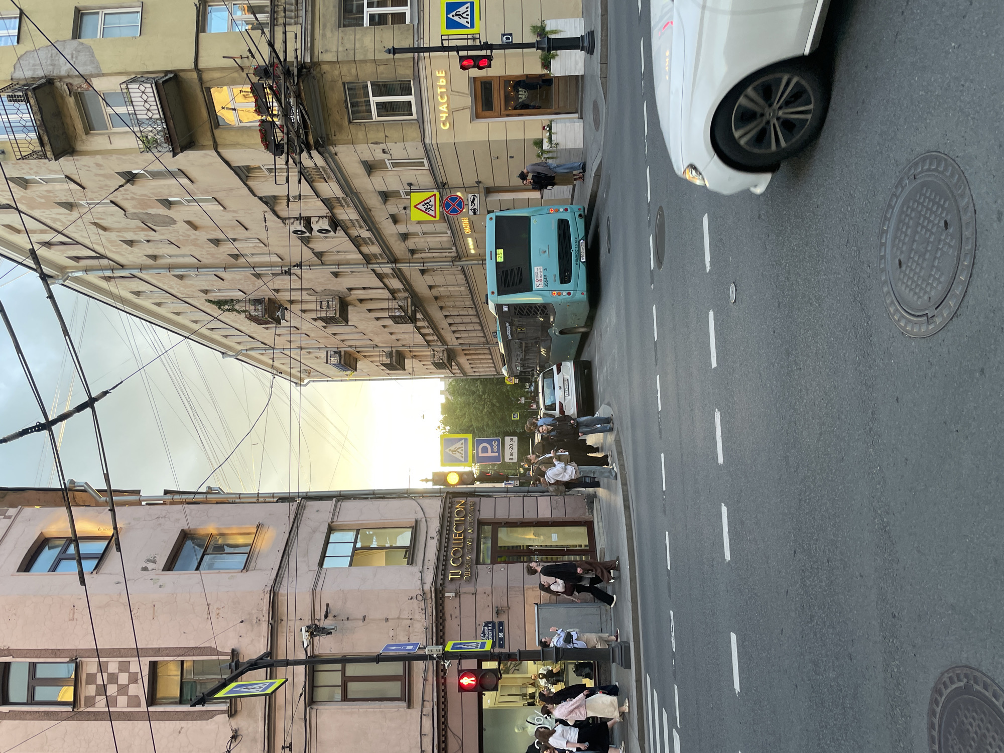 The image depicts a street scene in Saint Petersburg, Russia, likely at an intersection. In the foreground, a white car is partially visible on the right side of the frame. The street features crosswalk markings and manhole covers.
On the left, a light pink building with multiple windows and some architectural details is visible. A pedestrian crossing sign with a blue pedestrian symbol is mounted on a pole. A red traffic light is also present. A shop with the sign "TJ COLLECTION" and Russian text is located at the base of this building. Several pedestrians are walking on the sidewalk.
On the right, a light yellow building with a distinct façade, balconies, and numerous windows lines the street. A sign with the Russian word "СЧАСТЬЕ" (meaning "happiness") is prominently displayed. Below this sign, there is an entrance and planters with flowers. Further down the street, a blue bus is stopped, its rear visible. Traffic lights and various street signs, including pedestrian crossing and parking signs, are present. The sky above is overcast with hints of sunset light. The buildings exhibit signs of age and weathering.
