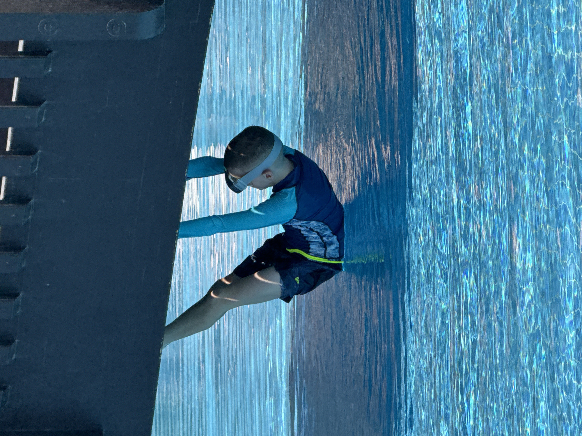 An upside-down, full shot shows a young boy with a snorkeling mask on his head, hanging from a dark, textured overhang above a bright blue swimming pool in Dubai, United Arab Emirates. The boy is wearing a long-sleeved blue rash guard and dark blue swim trunks with a neon green drawstring. His legs are bent at the knees and his feet are submerged in the pool water. The water's surface is rippled, reflecting the overhead overhang and creating a shimmering effect. The pool appears to be outdoors, with sunlight illuminating the water.