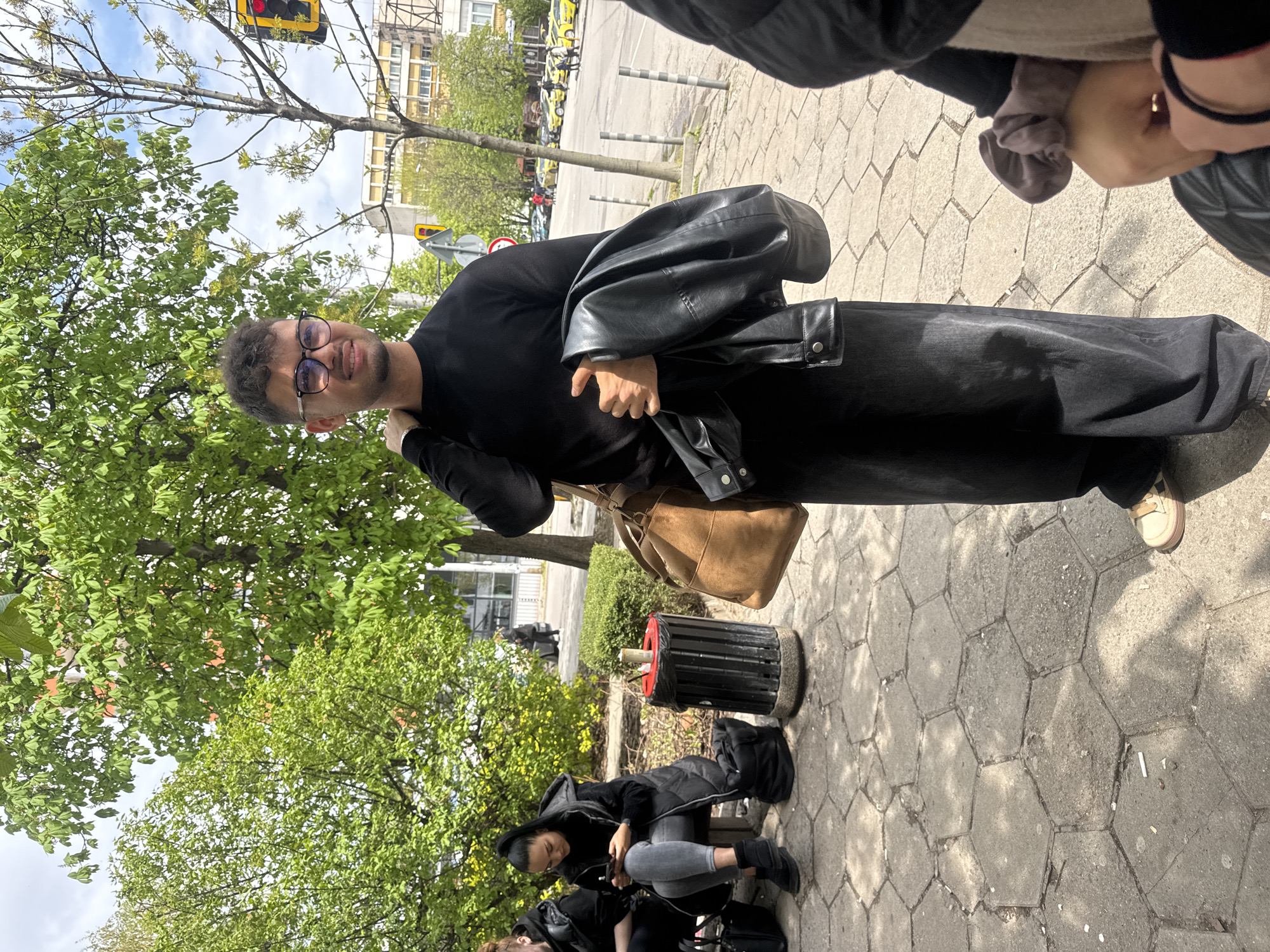 The image captures a man standing on a cobblestone pavement in what appears to be an urban setting. He is wearing a black long-sleeved shirt, wide-legged black trousers, and glasses. He has a black leather jacket slung over his left shoulder and a tan-colored bag hanging on his right side.
In the background, there are trees with green foliage, and in the distance, buildings are visible. A street with parked vehicles can be seen further away, with yellow taxis being identifiable. There's a traffic light hanging from a pole to the left of the man.
Closer to the foreground, a black cylindrical trash can with a red top sits near a bench where two individuals are seated. One of them is looking at a mobile phone. Parts of other people's bodies are visible in the upper right corner of the image, suggesting the photographer was part of a group. The lighting suggests it is daytime.
