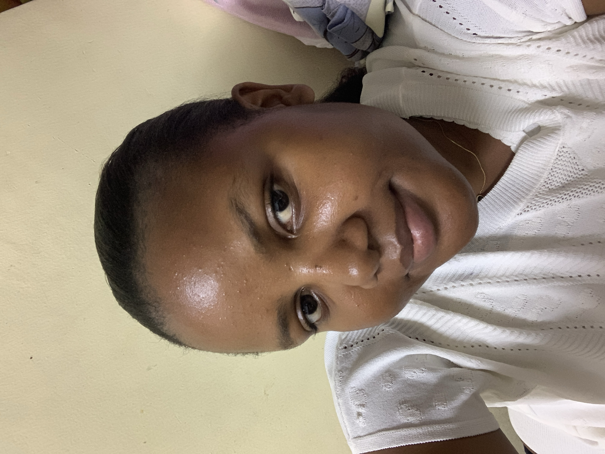 The image is a close-up selfie of a dark-skinned woman with short, black hair pulled back from her face. Her skin appears slightly oily. She is wearing a white, knitted top. A thin gold chain is visible around her neck. The background is a pale yellow wall. There is no information to ascertain if the photo was taken in Dar es-Salaam, Tanzania.