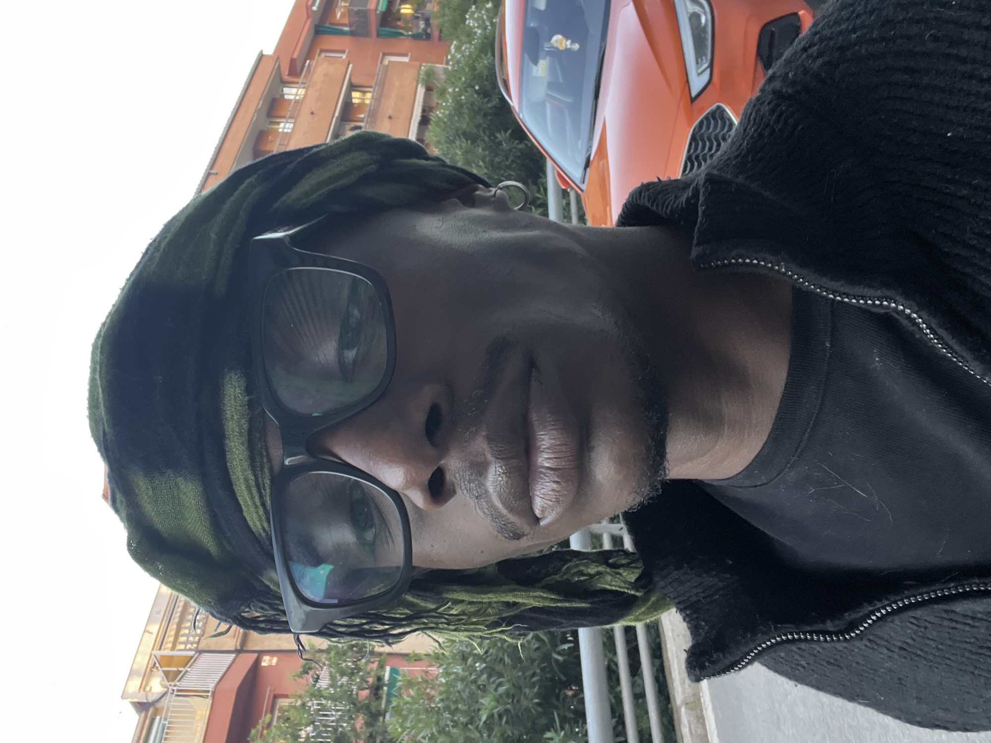 A close-up portrait depicts a dark-skinned individual wearing black-framed glasses and a green and black headscarf. The person is wearing a black t-shirt and a zippered black jacket. Behind the subject, there is a bright orange car and an apartment building with balconies. Green trees are also visible. The location is likely in Barcelona, Spain, based on the architecture of the buildings.