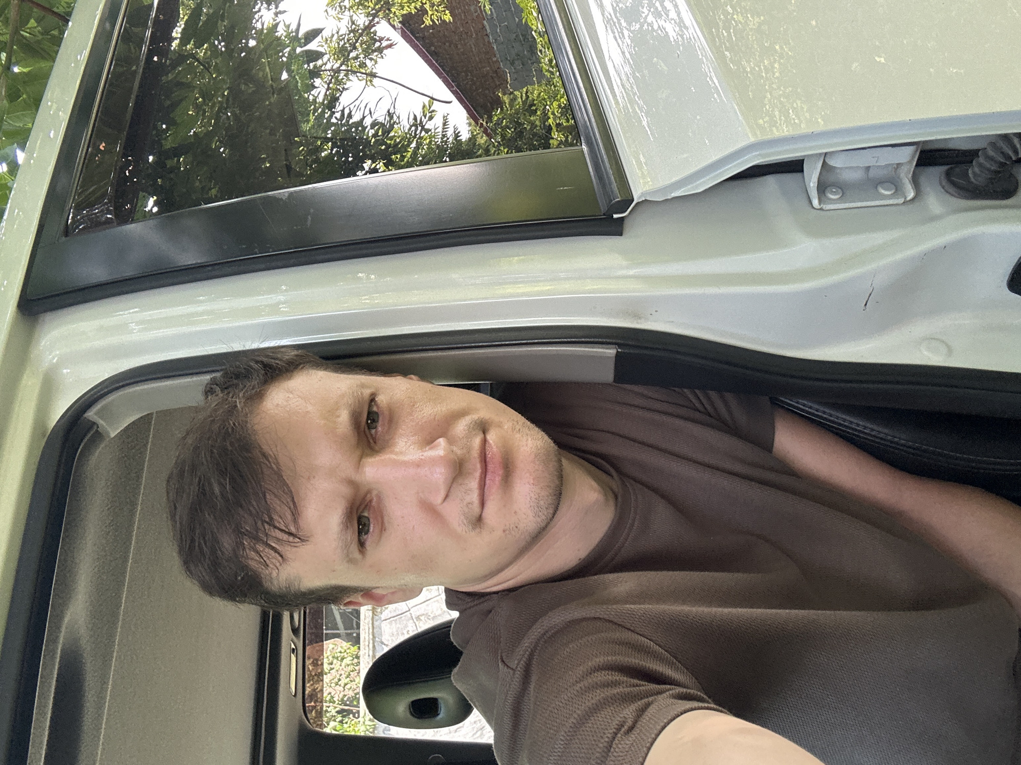 The image shows a close-up, selfie-style shot of a man sitting in the driver's seat of a car. He is looking directly at the camera with a neutral expression. The car door is open, revealing the exterior. The man is wearing a brown t-shirt. The interior of the car shows a headrest and part of the car's roof. Outside the car, through the open door and window, trees and a brick building are visible, suggesting an outdoor, possibly residential, setting. The sunlight indicates it is daytime. The location is Makiv, Ukraine.