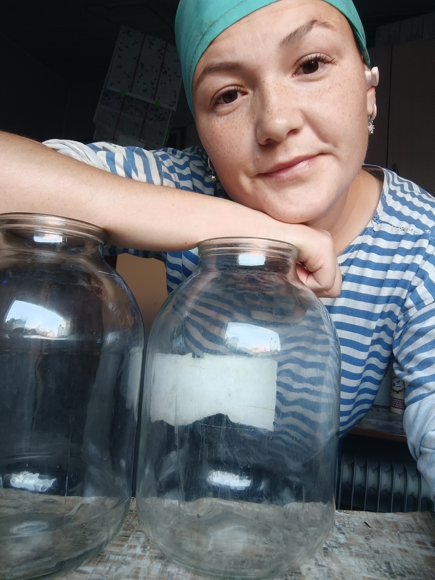 A young woman with freckles and wearing a teal headscarf and a blue and white striped shirt, is leaning her arm on two large glass jars. Her head is tilted, and she is looking towards the camera with a slight smile. One jar is to her left and is dark with reflections. The other jar is to her right and has a white label on it, with some contents visible inside. The background is dark, with some boxes stacked in the upper left corner. The scene appears to be indoors, possibly in a home. The location is not specified as Karsha, Russia.