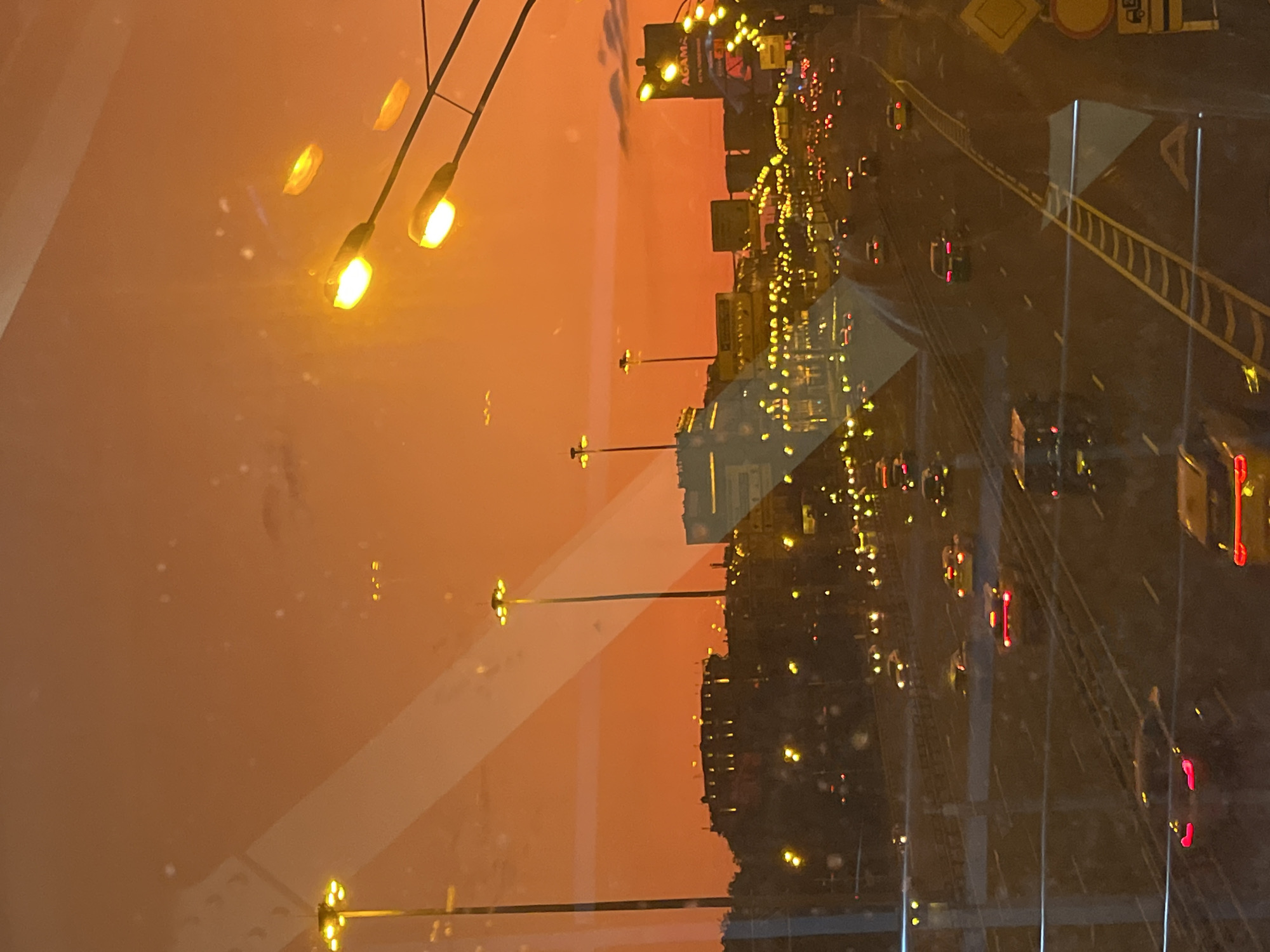 This is a photograph taken from a window overlooking a highway in Khimki, Russia, during a sunset with an orange sky. The highway is multi-lane and filled with cars, their headlights and taillights creating streaks of light. Several tall streetlights with bright yellow lamps are visible along the sides of the highway. In the background, there are buildings and structures, some illuminated by city lights. The image captures a typical evening scene of urban traffic and infrastructure against a dramatic sky.