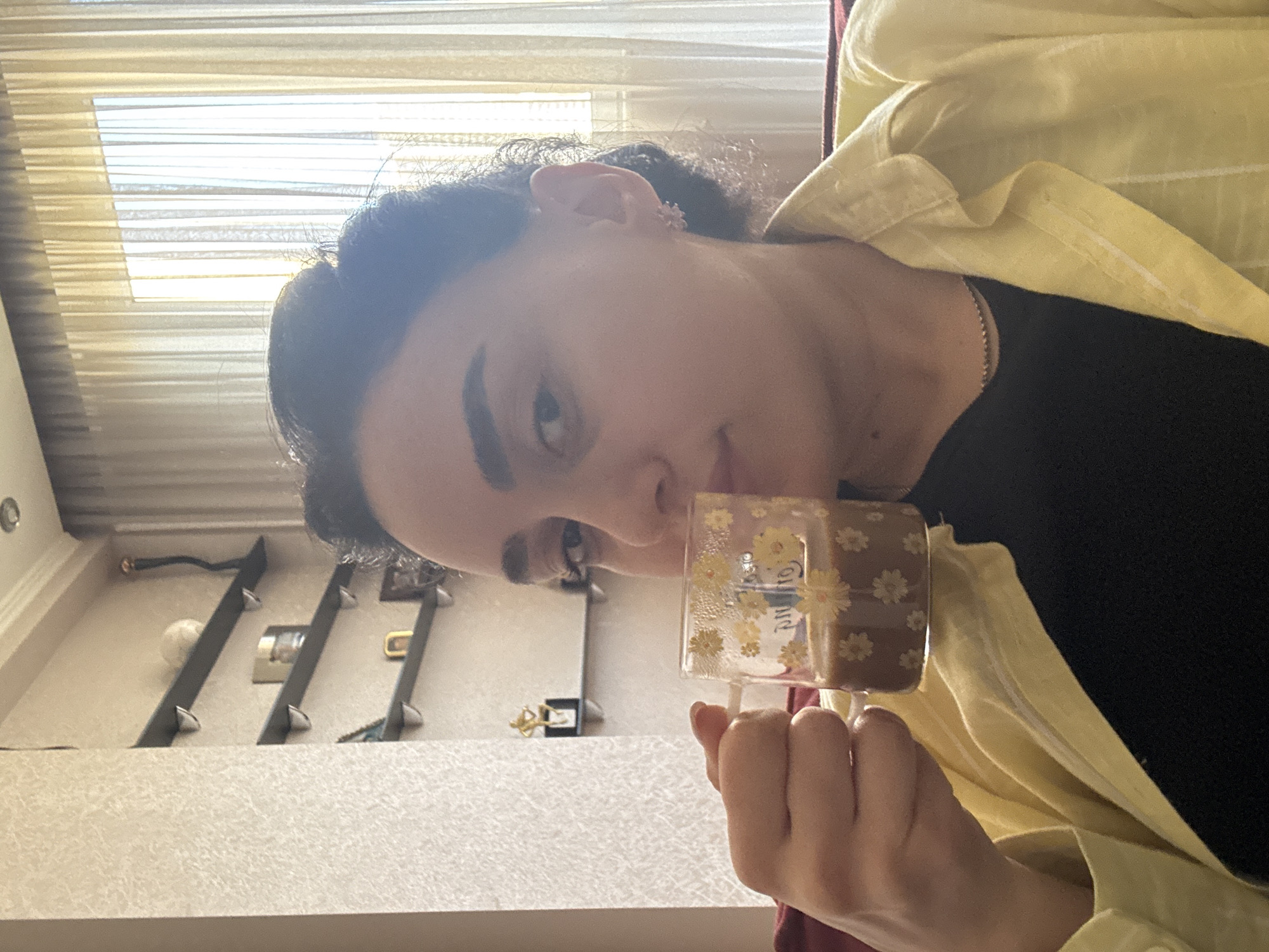 A woman is holding a mug with a floral design. She is wearing a yellow collared shirt over a black shirt. The background shows a window with blinds and shelves with various objects. The location is possibly an indoor space in Tbilisi, Georgia.