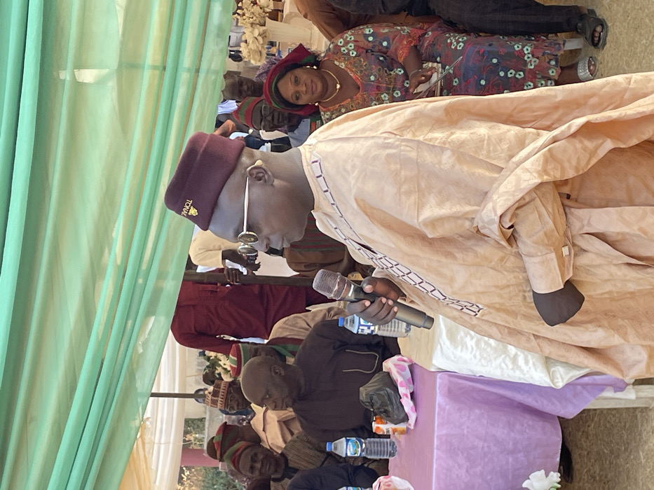 The image captures a social gathering or event, likely outdoors or in a semi-open space under a tent, indicated by the sheer light green drapes visible on the left side of the frame. The perspective is slightly elevated, looking down at the main subject and the audience.
In the foreground, a man is actively speaking into a microphone. He is dressed in a light-colored traditional garment, possibly a cream or beige agbada, with a patterned design visible along the trim. He wears a maroon or dark red cap that has golden text on its side, which appears to read "TONHA" or a similar word, possibly a name or an acronym. He also wears glasses with slender, light-colored frames and dark lenses. His right hand holds a black and silver microphone, while his left arm is partly visible, bent at the elbow.
In front of the speaker, a table draped with a lavender or light purple tablecloth is partially visible. On the table, there are several plastic bottles of water, some with blue caps, suggesting refreshments for attendees. Also on the table are some items in small plastic bags.
Behind the speaking man, and slightly to his right (from the viewer's perspective), a woman is seated. She is looking directly towards the camera with a gentle smile. She is wearing a vibrant, patterned dress in shades of red, blue, and brown, along with a matching headwrap. She accessorizes with gold-colored jewelry, including a necklace and earrings.
Further in the background and to the left of the main speaker, several other individuals are seated, forming part of the audience. Many are also dressed in traditional attire, including various caps, some striped and others solid-colored. One man in a dark brown or black garment is seated directly opposite the table, appearing to look downwards. Another individual in a maroon traditional outfit is also visible further back.
The overall setting suggests a formal or semi-formal event, possibly a celebration, meeting, or ceremony, given the speaker, microphone, and decorated environment with white floral arrangements visible in the far background. The lighting appears bright and natural, indicating it is daytime. The attire of the individuals suggests a cultural context, possibly West African.