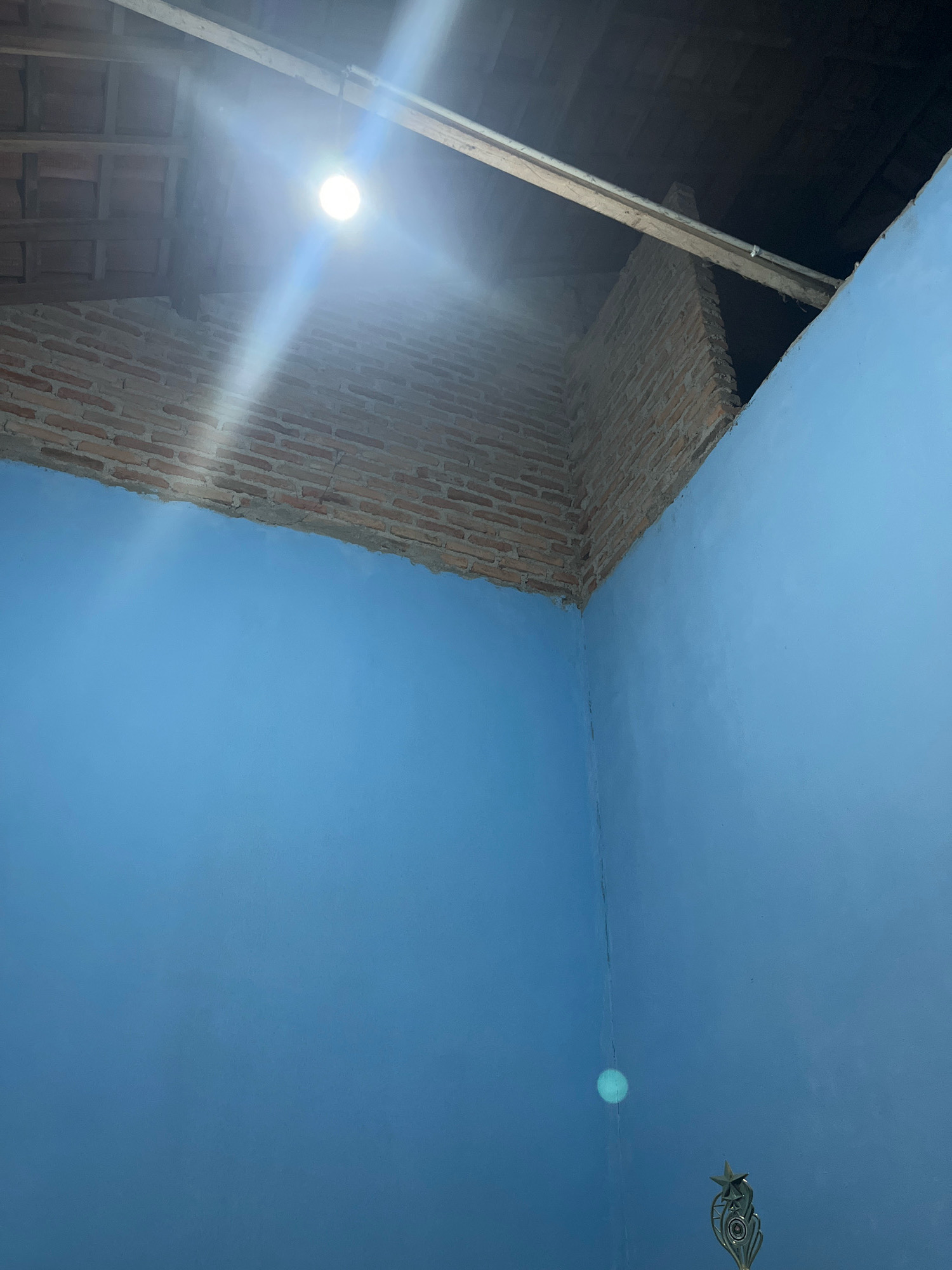 This image captures an interior corner of a room with unfinished brickwork visible at the top, suggesting a construction or renovation phase. The walls are painted a vibrant blue. A bright light bulb hangs from the ceiling, illuminating the scene and casting glare. A wooden beam runs across the upper section, supported by the brickwork. The flooring is not visible. The image is taken from a low-angle perspective, looking up towards the ceiling. The room appears to be located in a residential area in Kebumen, Indonesia.