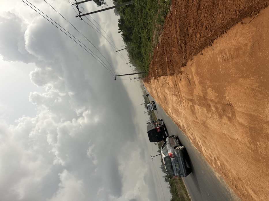 The image depicts a rural or semi-urban roadway in Ilishan, Nigeria, under an overcast sky filled with dense grey and white clouds. The scene is dominated by an asphalt road running diagonally from the lower-left foreground towards the center-right horizon. Adjacent to the right side of the asphalt, a wide section of recently disturbed red-brown earth forms an unpaved shoulder or a construction area. This dirt varies in texture, suggesting recent grading or excavation.
On the asphalt road, multiple vehicles are visible, all moving away from the viewer. In the immediate foreground, a grey station wagon or hatchback with a visible license plate frame is present. Behind it, a dark-colored panel van or large SUV proceeds. Further down the road, a white SUV is observed, followed by a distant motorcycle.
To the right of the dirt shoulder, a series of utility poles with overhead power lines extends parallel to the road, disappearing into the distant haze. Beyond these poles and the dirt area, green vegetation, including trees and bushes, lines the roadside. No pedestrians are visible.