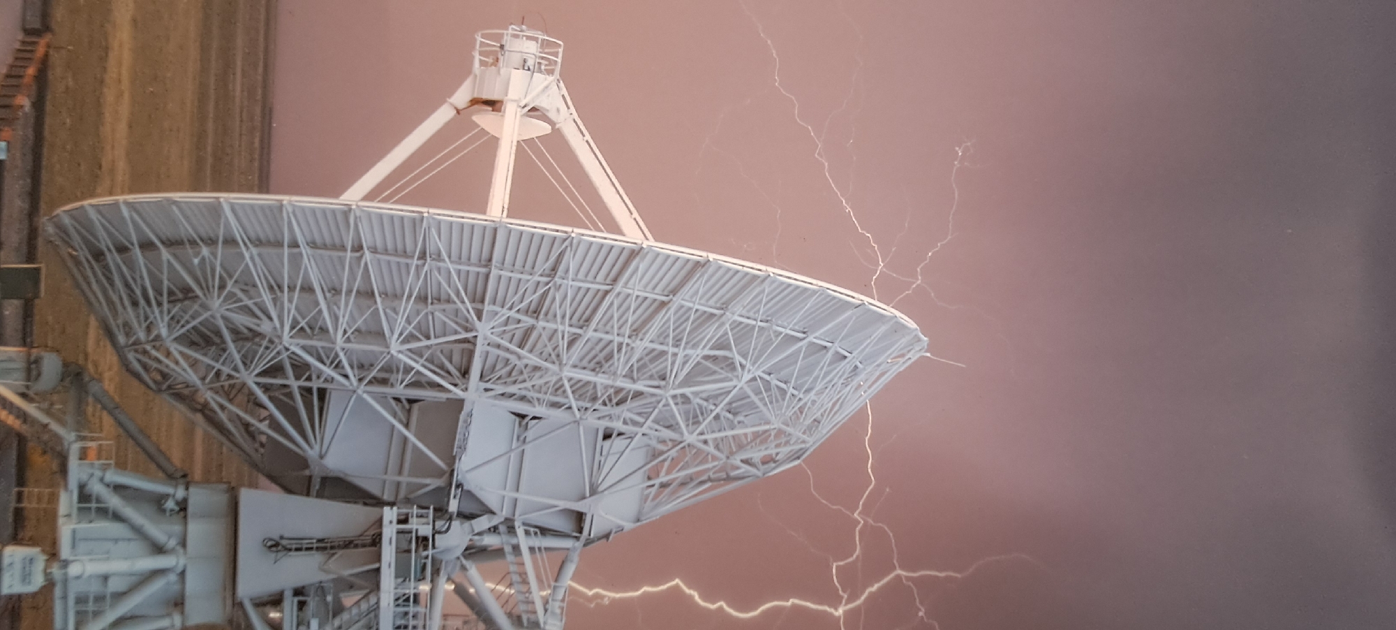 The image shows a large, white, parabolic satellite dish with a complex lattice structure. The dish is positioned at an angle, with its reflective surface facing towards the left of the frame. Behind the satellite dish, the sky is a dusky pinkish-brown, with numerous white, branching lightning bolts illuminating the scene. The lightning appears to be a significant storm in progress. In the foreground, the base of the satellite dish is supported by a sturdy, white metal structure, and the ground beneath it is a dry, tan-colored plain or desert. The overall scene evokes a sense of power and drama, with the artificial structure of the satellite dish contrasting with the natural phenomenon of the lightning storm. The image does not provide any visual cues to suggest a location in Hässelby, Sweden.