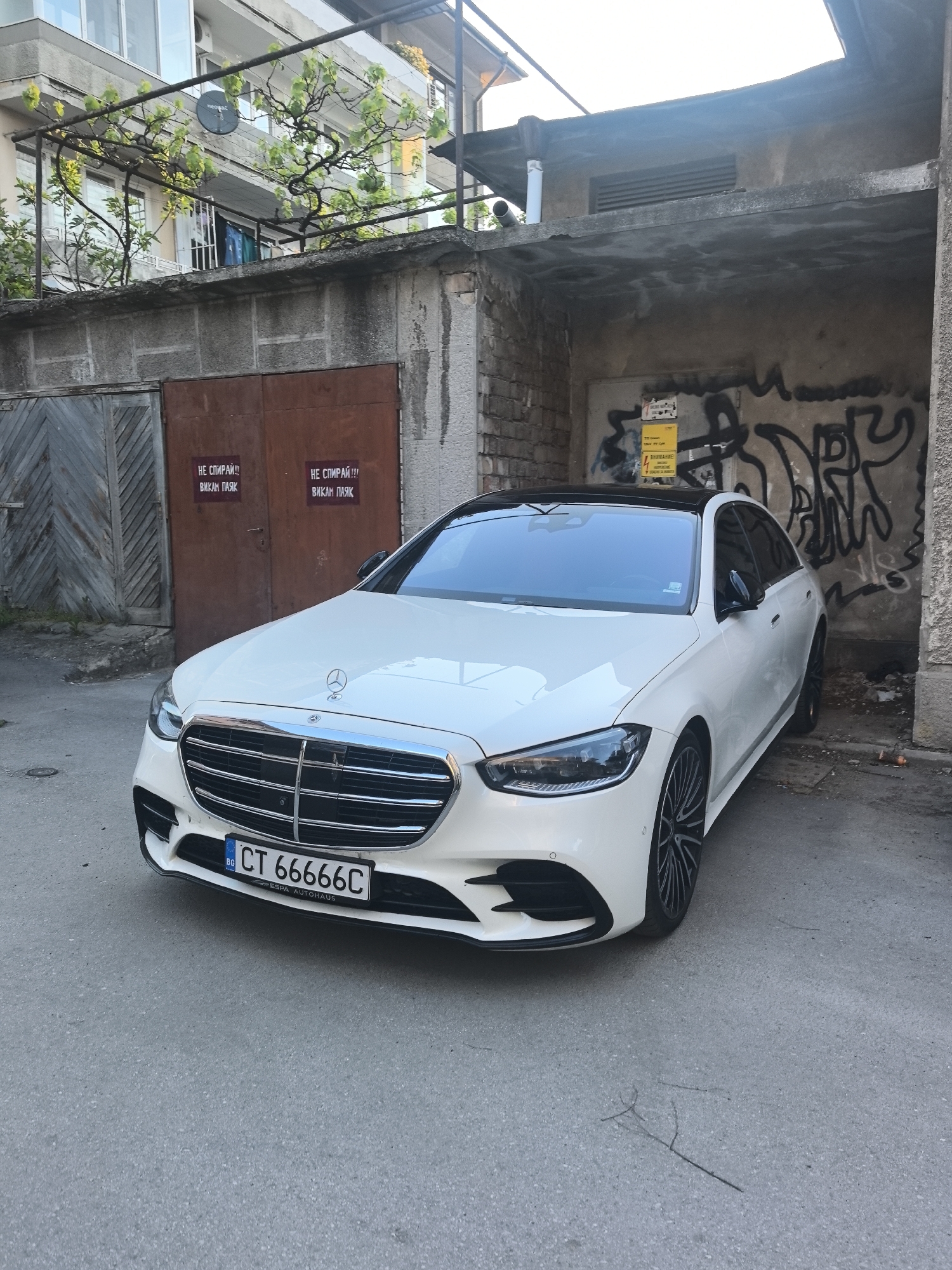 The image depicts a white Mercedes-Benz S-Class parked on an urban street in Stara Zagora, Bulgaria. The car has a black roof, chrome grille, and a Bulgarian license plate reading "CT 66666C".
The car is positioned in front of a concrete structure with visible graffiti on the wall to the right of the car. A yellow warning sign is attached to the wall with graffiti behind the car. There are metal doors to the left of the car with posters adhered to them. The posters have text written in Bulgarian. There are plants and a building in the background on the left. The street is paved and appears to be in a residential or commercial area.