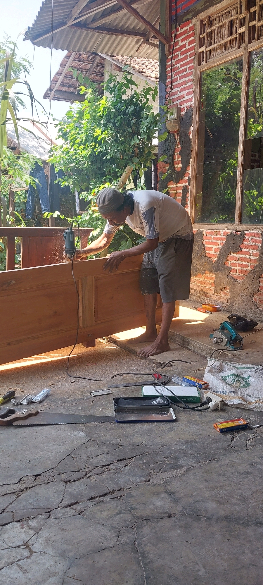 A man is working on a wooden door panel outdoors. He is using a router tool, held in his right hand, to shape the edge of the wood. He is wearing a grey cap, a light-colored t-shirt, and dark shorts. The wooden panel is large and appears to be part of a door frame. Tools and materials, including a saw, a planer, and several boxes, are scattered on the concrete floor. The scene is set against a backdrop of a building with a corrugated roof and a brick wall, and lush green foliage. This location appears to be in Keling, Indonesia, as indicated by the context. The overall impression is of a woodworking activity taking place in a semi-outdoor workshop.