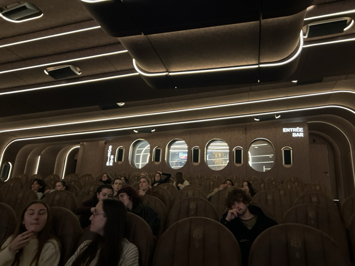 A movie theater with brown leather seats is partially filled with viewers. The theater features an "Entrée Bar" sign and porthole-like windows revealing shelves of refreshments. Striped lighting accentuates the curved architecture of the ceiling and walls. Some patrons are visible in the foreground, appearing relaxed or engaged.