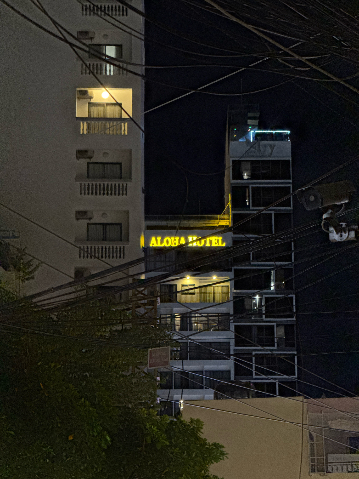 The image captures a nighttime street scene in Nha Trang, Vietnam, dominated by the illuminated sign of "ALOHA HOTEL" in yellow neon. The hotel building is tall and modern, with multiple balconies and windows. Another, taller building stands to the right, also with a modern facade, featuring a row of windows that appear recessed. A complex network of overhead electrical wires crisscrosses the frame, obscuring parts of the buildings and the night sky. A security camera is visible mounted on one of the poles supporting the wires. In the foreground, a leafy tree partially obscures a red sign with white text, which is difficult to read due to its distance and the low light conditions. The overall atmosphere is urban and somewhat cluttered due to the dense wiring.