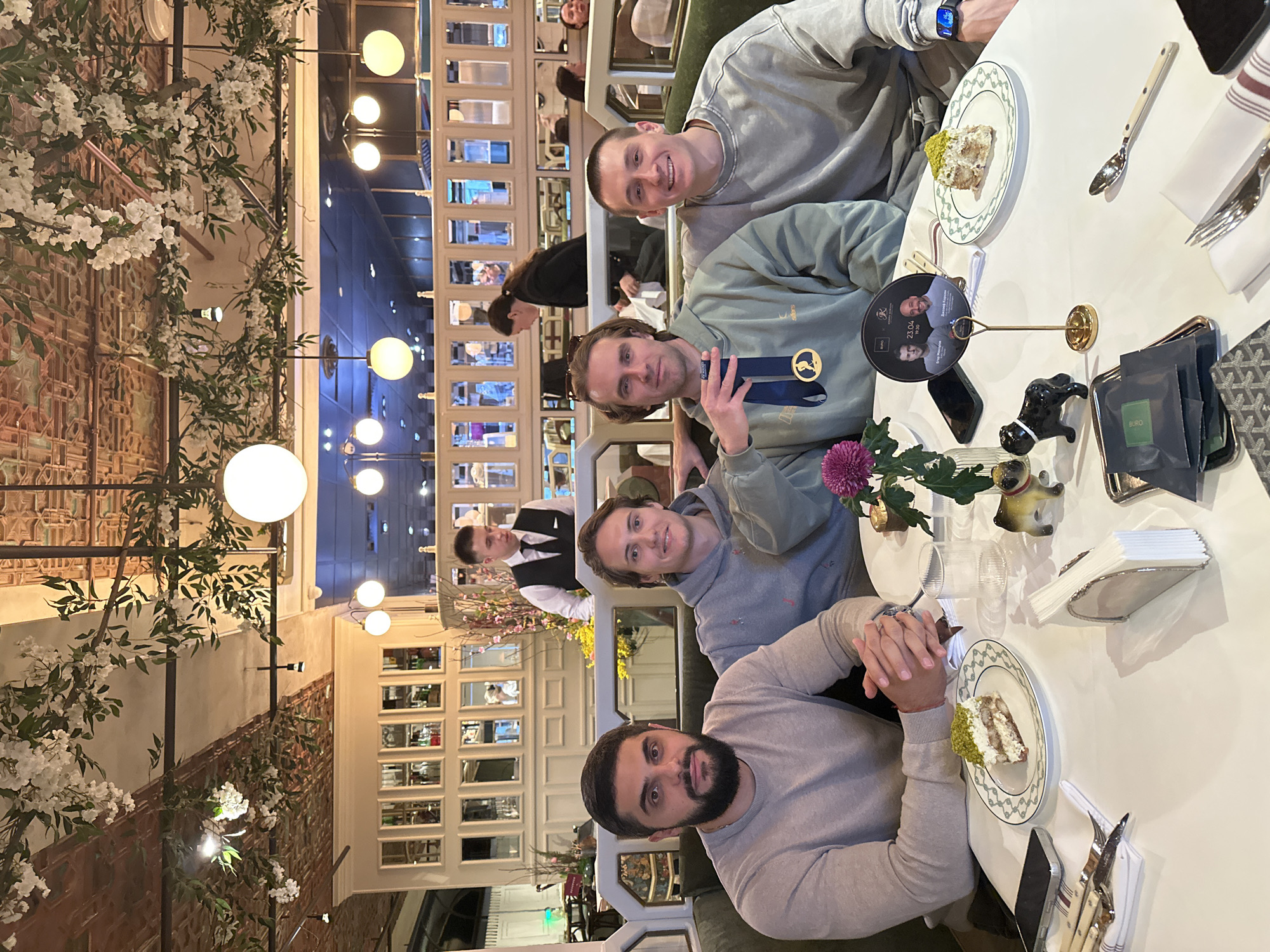 Four men are seated at a white table in a restaurant. Each man is wearing casual clothing. One man is holding a blue ribbon or medal. There are plates with slices of cake on the table. A flower vase and a small dog figurine are also visible. A waiter stands in the background. The restaurant has an upscale appearance with decorative floral arrangements and mirrored walls. The establishment is Buro TSUM Restaurant, located in the city of Moscow, Russia.