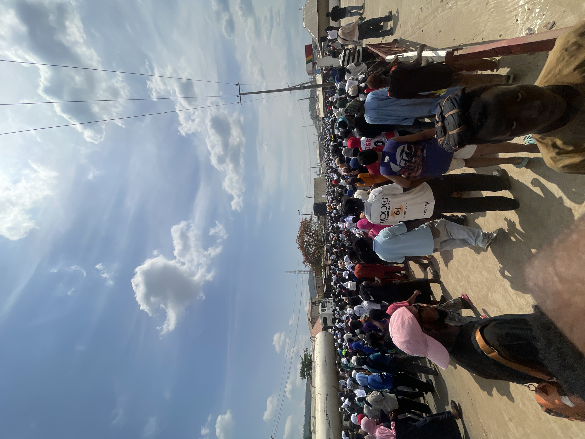 A large group of people are gathered in an outdoor area in Lokoja, Nigeria. Many individuals are wearing white clothing and head coverings. A fuel tanker with the letters "OPL" can be seen on its side, as well as a tree with reddish-orange leaves. Overhead power lines are visible against a blue sky with scattered clouds.