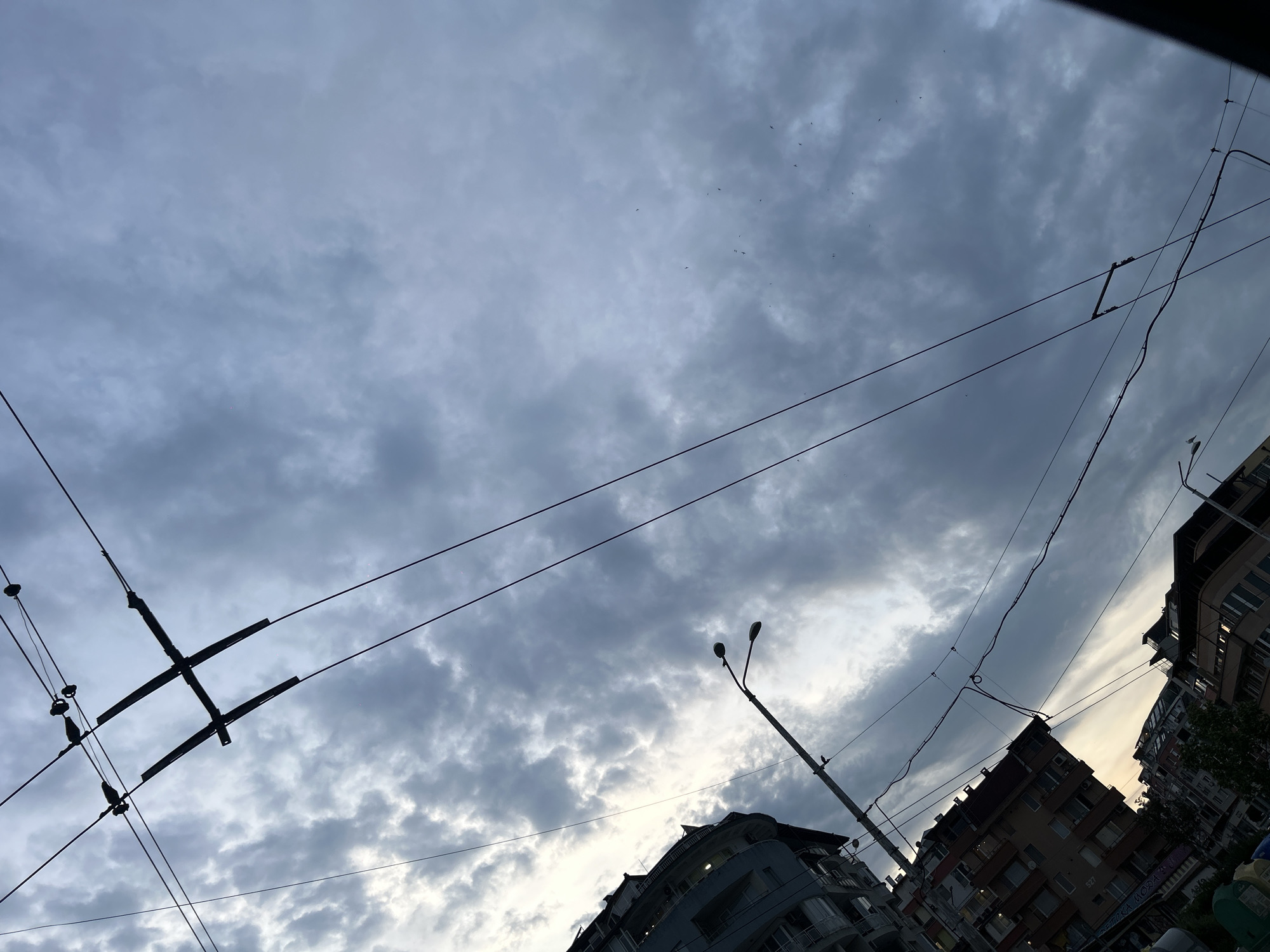 The image depicts a low-angle view in an urban environment, likely in Burgas, Bulgaria, based on architectural style and urban infrastructure. Predominantly, the view is of an overcast sky with layered, greyish-white clouds. A group of small birds is visible in the sky. Power lines are stretched across the frame, intersecting at points, and a light pole extends upward. Parts of buildings are seen at the bottom, including balconies, windows, and architectural details typical of urban construction. The lighting suggests either dawn or dusk, with the sky being the primary light source. The overall impression is of a typical street scene in an urban area with a cloudy sky.