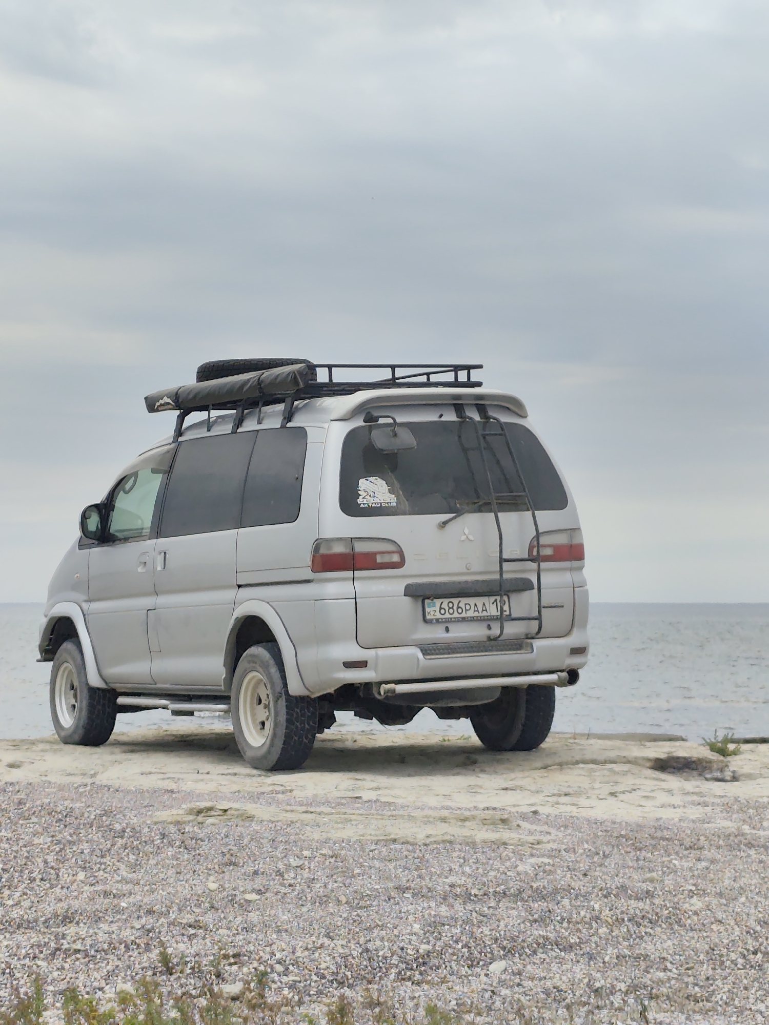 A silver Mitsubishi Delica van, equipped with a roof rack carrying a spare tire and an awning, is parked on a rocky shore next to the Caspian Sea in Aqshuqyr, Kazakhstan. The van has off-road tires and a ladder mounted on the rear. The license plate is visible and reads "KZ 686PAA 12". The sky is overcast. The environment is natural, with the sea in the background and a gravelly beach in the foreground. There are no people visible in the image.