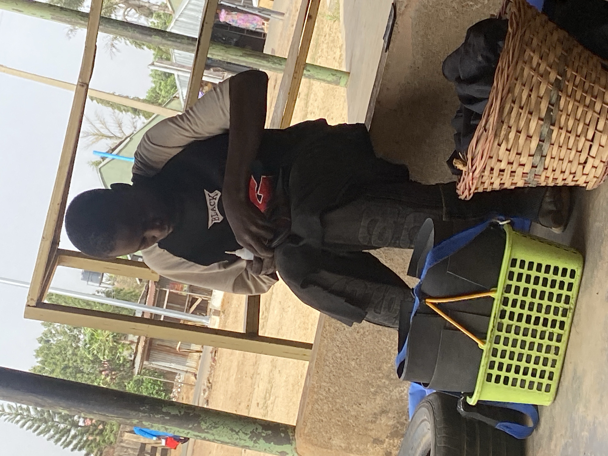 A person wearing a black t-shirt with white lettering and a red symbol is seated on a concrete surface. The person is bent over, looking down. Adjacent to the person are a brown wicker basket filled with dark clothing, a lime green plastic crate containing black items secured with yellow straps and blue straps, and a tire. The location appears to be outdoors, with a wooden structure overhead. Background details suggest the city of Jos, Nigeria.