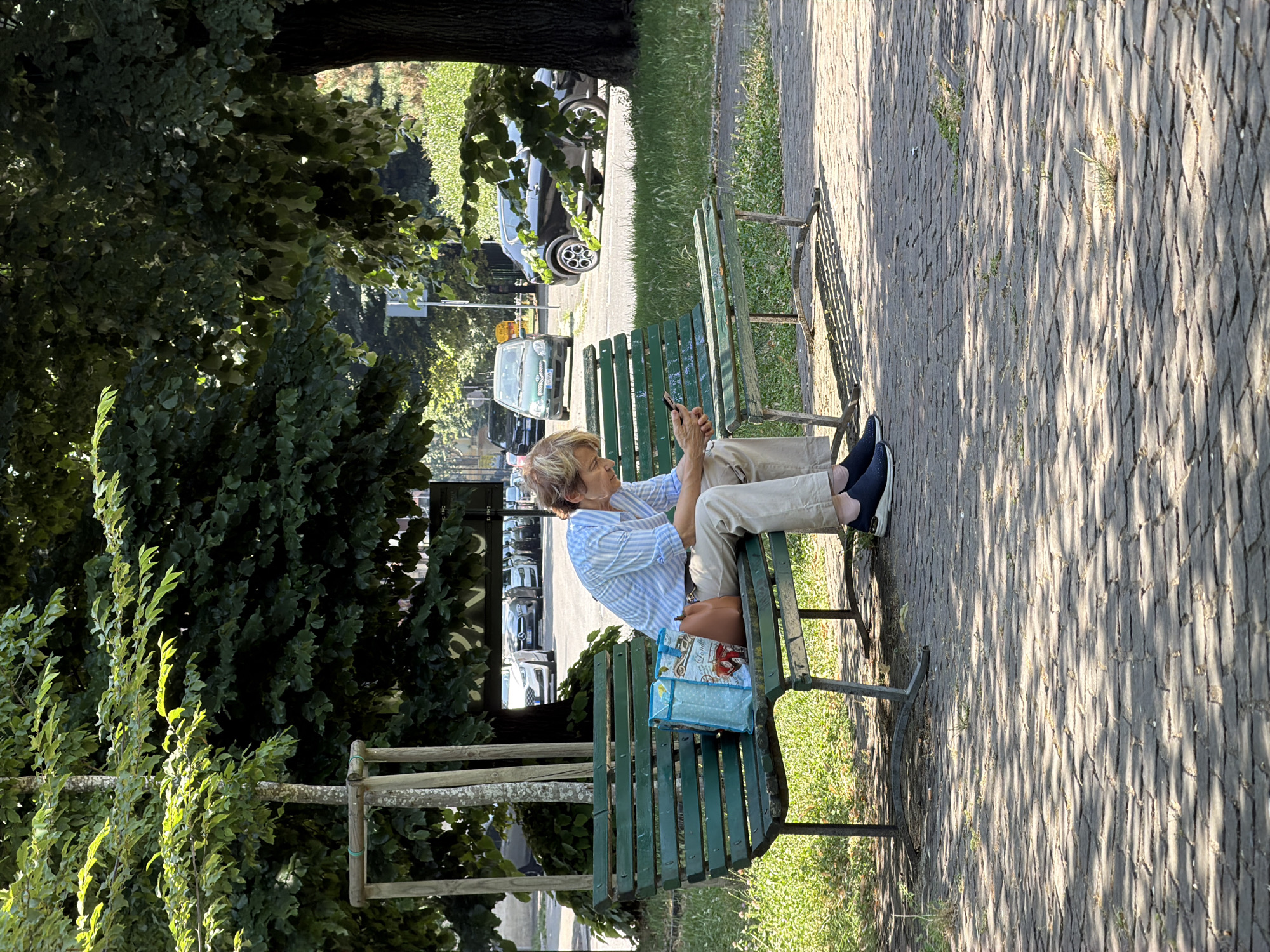 In Milan, Italy, a person is seated on a green park bench, engrossed in their smartphone. The individual, with blonde hair, is wearing a striped blue and white button-up shirt and light-colored pants, along with dark shoes. Beside them on the bench are two bags: a blue and white patterned tote bag and a brown handbag. The bench is positioned on a paved pathway, surrounded by green grass and lush trees that cast dappled shadows on the ground. In the background, a street with several cars is visible, suggesting an urban park setting. The lighting indicates it is daytime, with the sun illuminating parts of the scene.