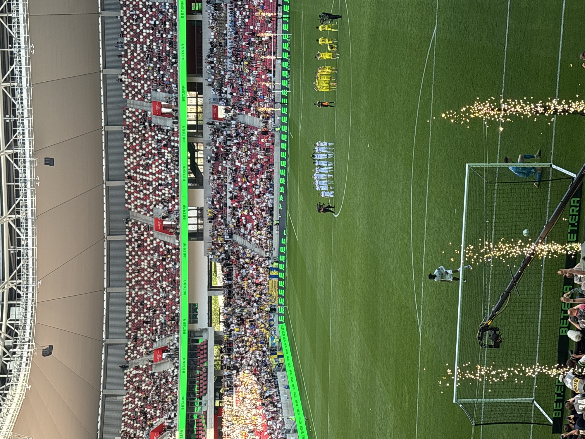 Here is a detailed description of the image:
The image presents a high-angle view of a soccer game taking place in a large stadium. The setting appears to be Minsk, Belarus, based on the "BETERA" signage visible in the image.
The stadium is filled with spectators, predominantly wearing red and white, suggesting they are fans of a particular team. The seating arrangement forms patterns of red and white throughout the stands.
On the green soccer field, two opposing teams are lined up, one in white uniforms and the other in yellow. The two teams are positioned at different ends of the field, with their respective coaches or managers standing nearby. A penalty kick is in progress, with a player in white uniform in position to kick the ball, as the goalkeeper dives to defend the goal. There are photographers around the goal, capturing the event. Special effects or lights are being used, possibly fireworks or sparklers, creating a sparkling effect surrounding the goal area.
The stadium structure includes a roof supported by metal frameworks. The image is taken during daylight, with natural light illuminating the scene. The image shows a blend of sports action, audience participation, and event-related spectacles.