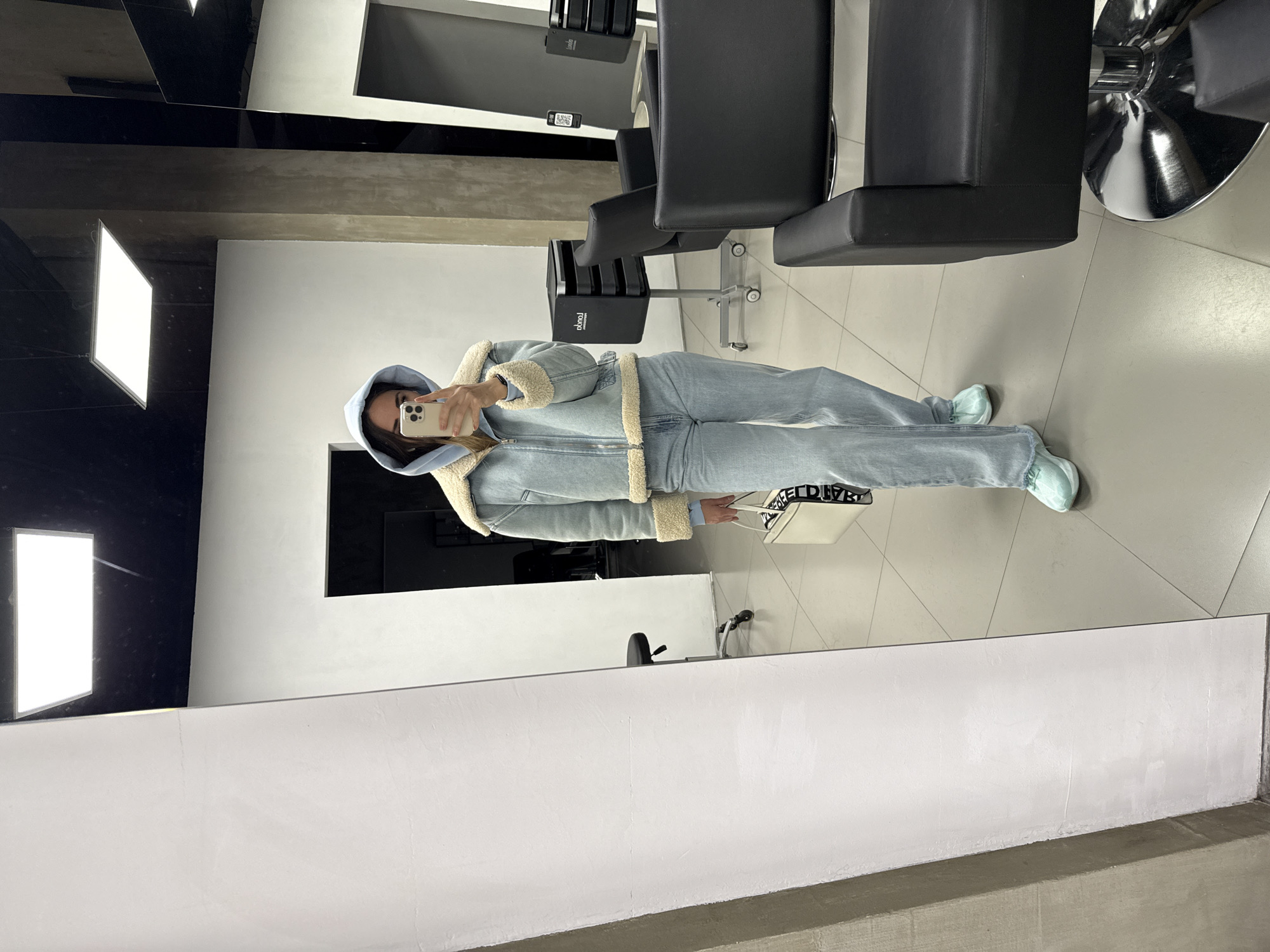 A person is taking a selfie in front of a mirror in what appears to be a hair salon or barber shop. The person is wearing a light blue denim jacket with a fur lining and matching jeans, and a blue hoodie under the jacket. The person is holding a smartphone in their right hand, angled towards their face.
In the background, there are several black salon chairs, a large mirror covering the back wall, and tiled flooring. Artificial lighting fixtures are visible on the ceiling. It cannot be definitively stated that the location is in Saint Petersburg, Russia, without additional clues or information.
