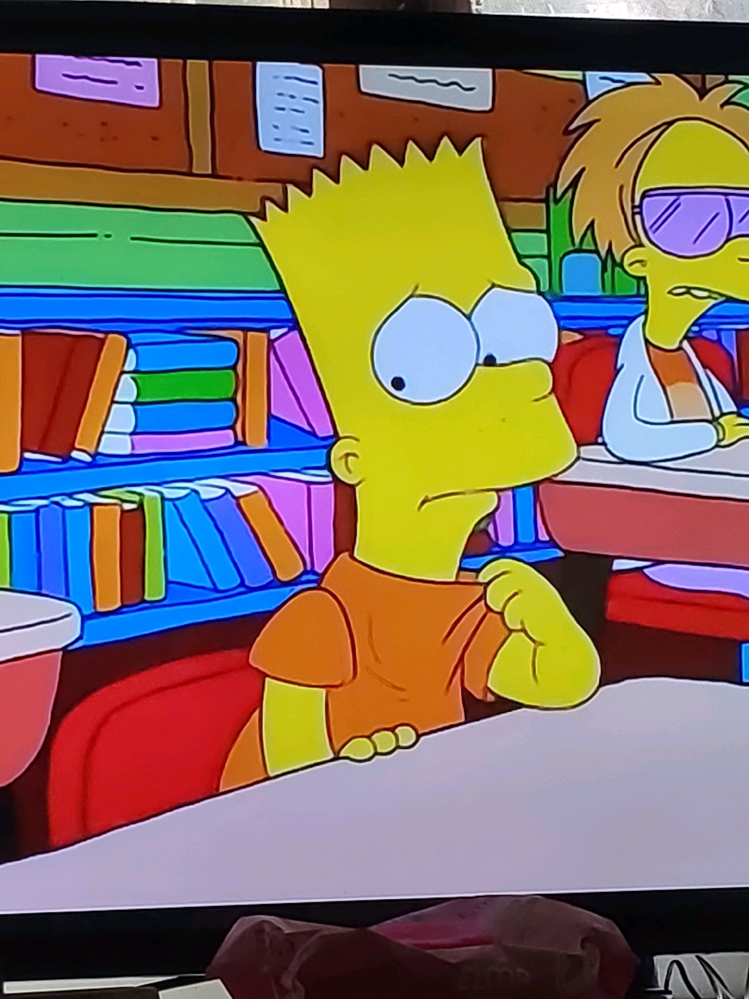 The image shows a scene from the animated television show The Simpsons. Bart Simpson, a young boy with spiky yellow hair and wearing an orange shirt, is seated at a desk in a classroom. His head is tilted and his eyes are wide with an apparent expression of concern or confusion. He is resting his chin on his hand. To his right, another student with blond hair and purple sunglasses is also visible, looking forward. The background of the classroom features bookshelves filled with colorful books and educational posters on the wall. The scene is depicted in the characteristic animation style of The Simpsons. There is no discernible information within the image that would associate it with Dustabad (Soldatsky), Uzbekistan, or any specific city. The image is a still frame from a television program.