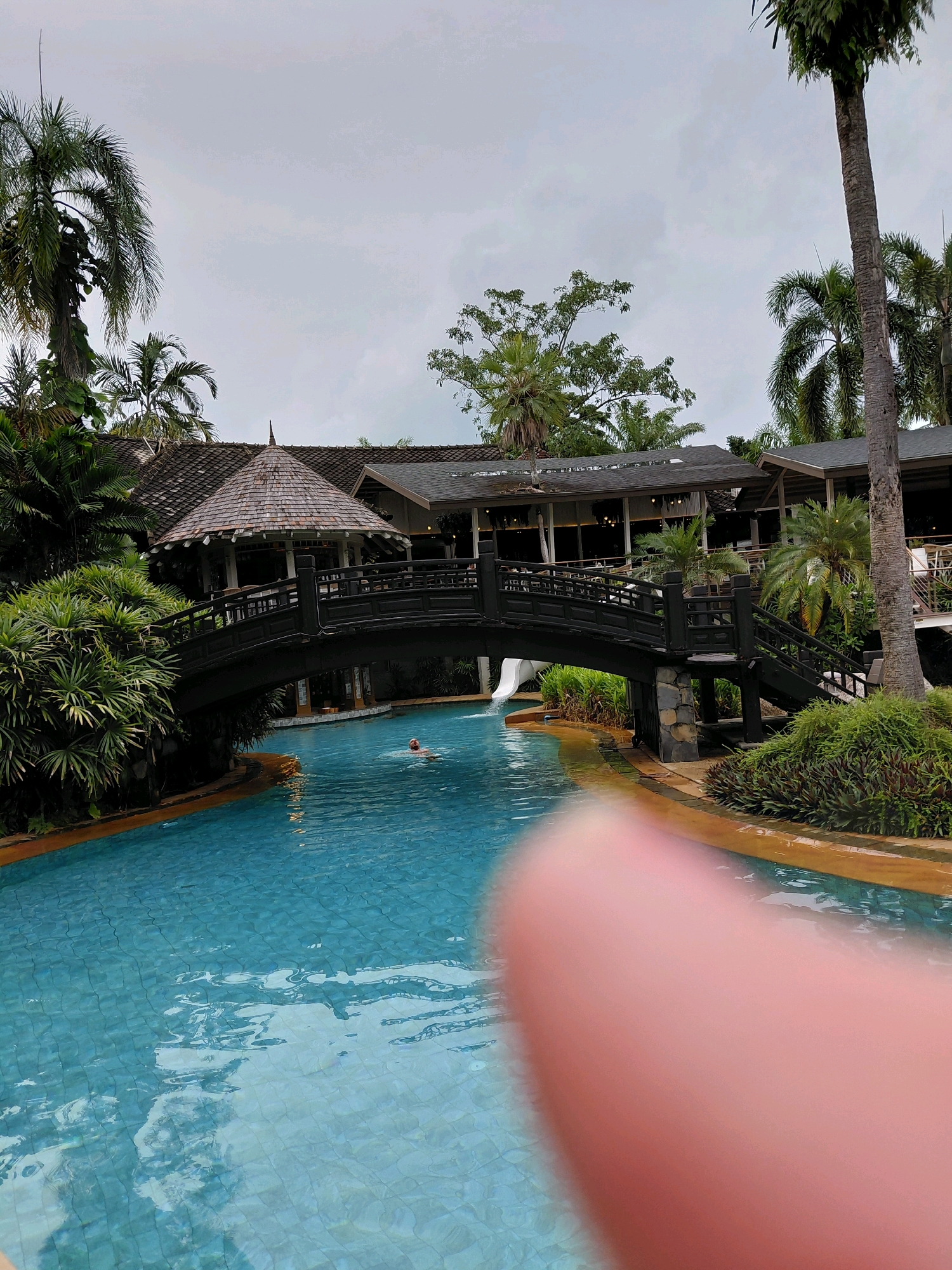 The image shows a swimming pool area in what appears to be a resort. A person is swimming in the bright blue water of the pool. A dark wooden bridge with decorative railings spans across the pool, connecting two buildings with traditional Thai architecture. A white water slide is visible on the left side of the bridge. Lush green tropical foliage and palm trees surround the pool and buildings. The sky is overcast. The overall scene suggests a vacation or resort setting. While the prompt mentions Ban Khuan, Thailand, this specific location cannot be confirmed solely from the visual information in the image.