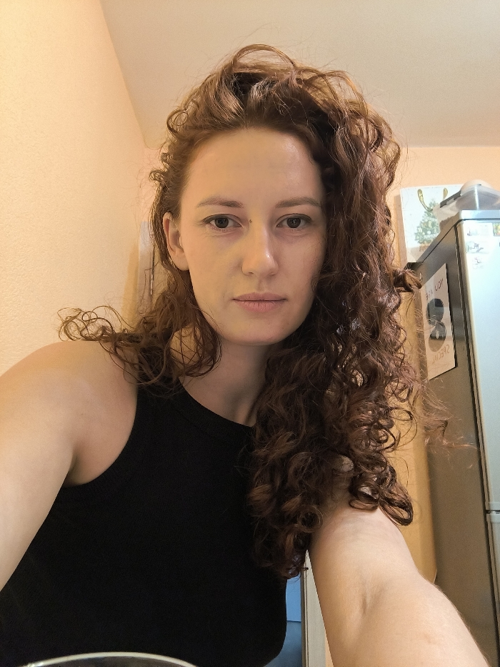 A headshot-style portrait captures a female subject from the chest up, positioned centrally. The subject has light skin, dark brown eyes, and long, heavily curled reddish-brown hair cascading over her shoulders. A small dark mole is present on her left cheekbone, inferior to her left eye. She wears a plain black sleeveless high-neck top. Her expression is neutral, and her gaze is directed straight into the camera lens. The background features a plain, light peach-colored wall on the left. To the right, a silver-colored two-door appliance, consistent with a household refrigerator, is visible. Two items are affixed to the refrigerator: an upper white rectangular magnet displaying a green graphic resembling a stylized Christmas tree or reindeer antlers, and a lower dark circular magnet with white text, partially revealing the word "SPECIAL." The environment is an interior space, consistent with a residential setting in Saint Petersburg, Russia. No specific address or architectural landmark within the city is discernible from the visual information provided.