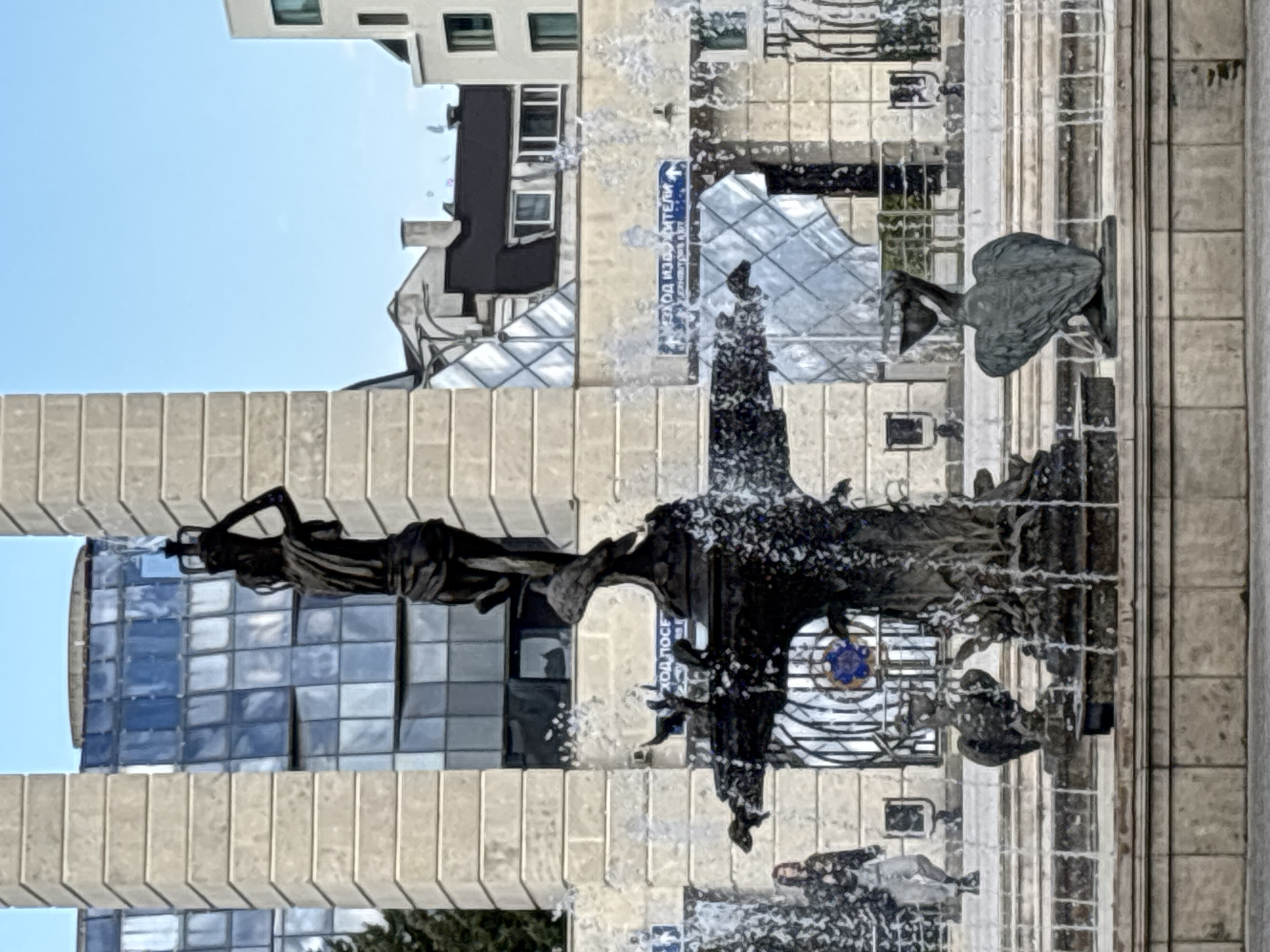 The image shows a fountain in Plovdiv, Bulgaria. The fountain has a sculpture of a woman standing atop a structure with multiple tiers. Water is spraying from various points in the fountain. In the background is a mix of modern buildings with reflective glass and older architecture. A sign with Cyrillic text is visible on the side of one of the buildings. There is also a sculpture of a bird, seemingly an eagle, in the background. The sky is light blue, suggesting a clear day.