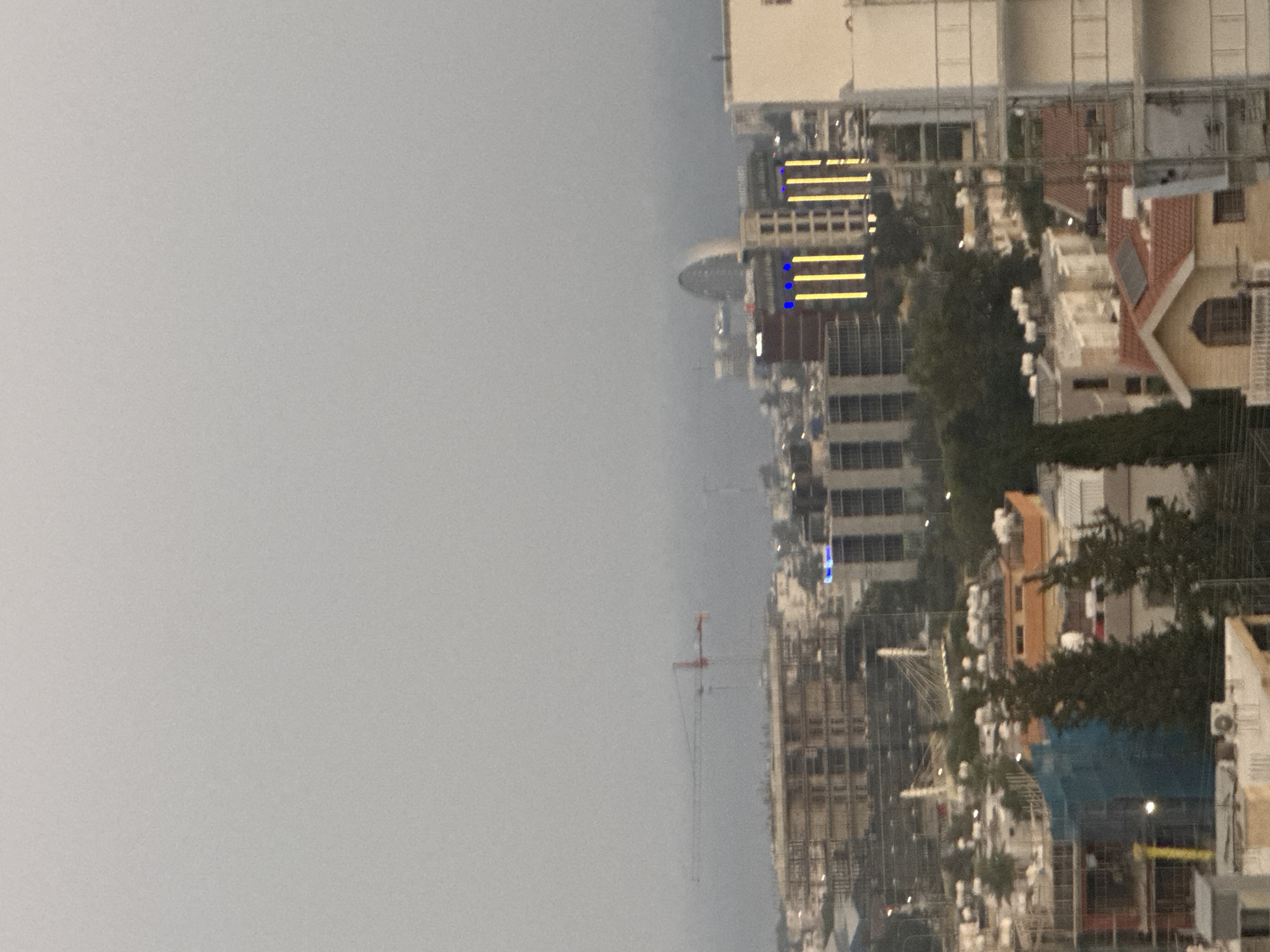 The image shows a city skyline with multiple buildings under construction and completed structures. In the background, a large red and white crane is visible, suggesting ongoing development. Several multi-story buildings are present, with some featuring illuminated vertical yellow lights. A distinctive arched building is also visible in the distance. The environment appears to be somewhat hazy or overcast. The scene is set in the Agios Athanasios Municipality, Cyprus.