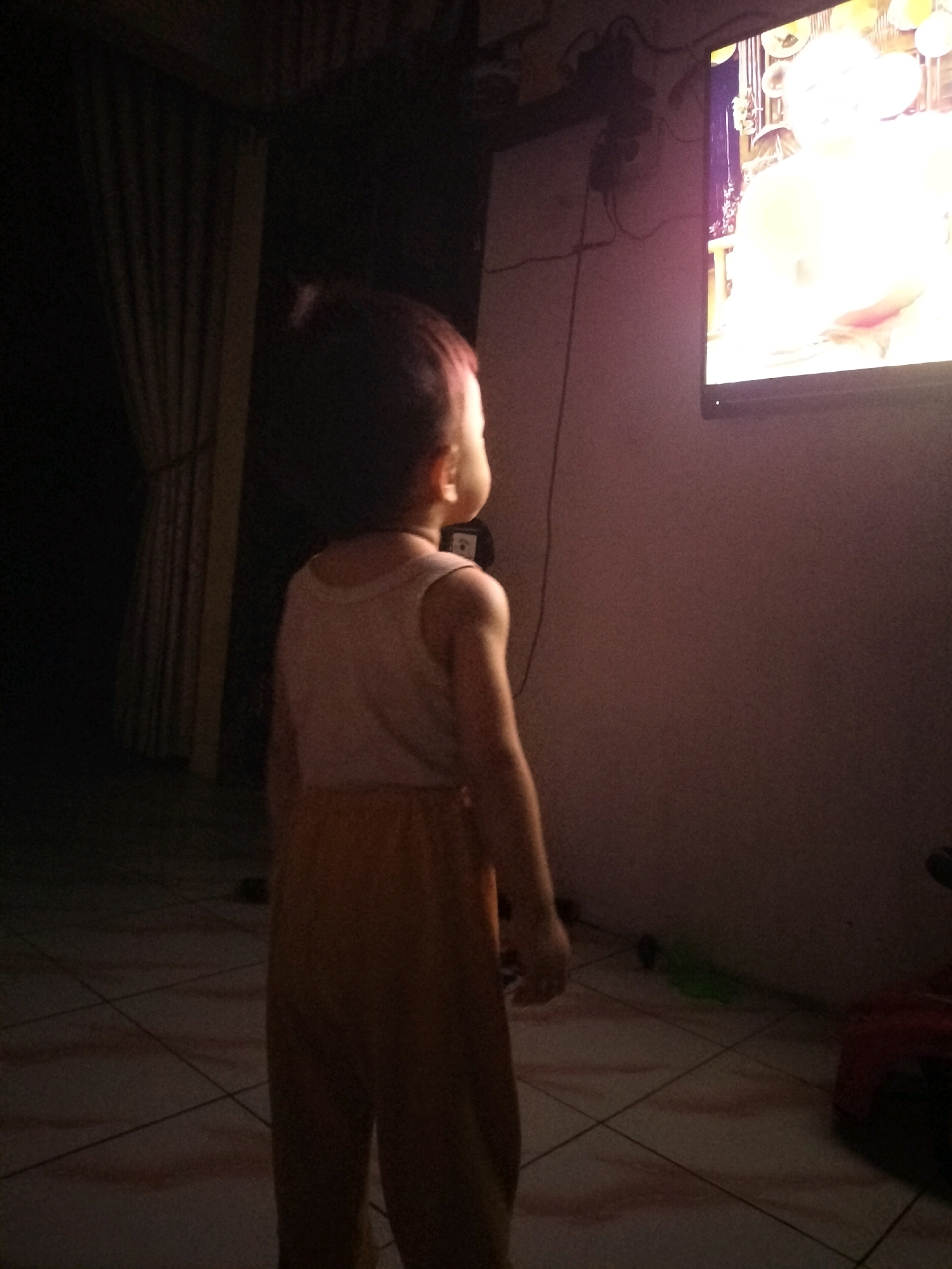 A young child stands in a dimly lit room, facing a television screen that is illuminated with a bright, colorful image. The child is wearing a light-colored sleeveless shirt and orange pants. Their back is to the camera, and their head is turned to the side, engrossed in what is on the television. The room has tiled flooring and a curtain hanging on the left side. The overall atmosphere is one of quiet observation, with the television providing the main source of light and focus. The time of day appears to be evening or night, given the darkness of the room.