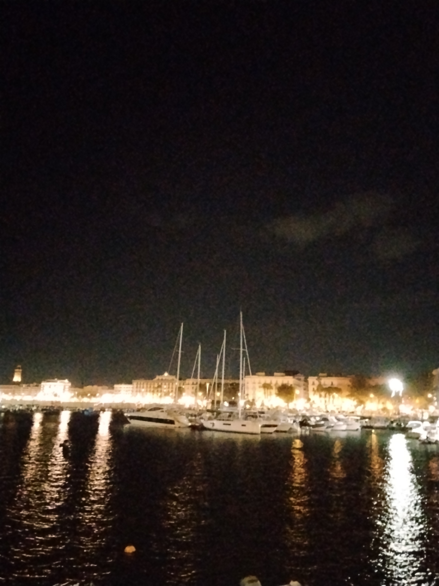 The image depicts a nighttime view of the port of Bari, Italy. Several sailboats and yachts are docked in the harbor, their white hulls and masts illuminated by the ambient city lights. The water reflects the lights of the city skyline, creating shimmering patterns. In the background, a city skyline is visible with buildings and a distant tower, all lit up by artificial lights. The sky is dark, with faint clouds. The overall scene conveys a sense of calm and tranquility associated with a port at night.