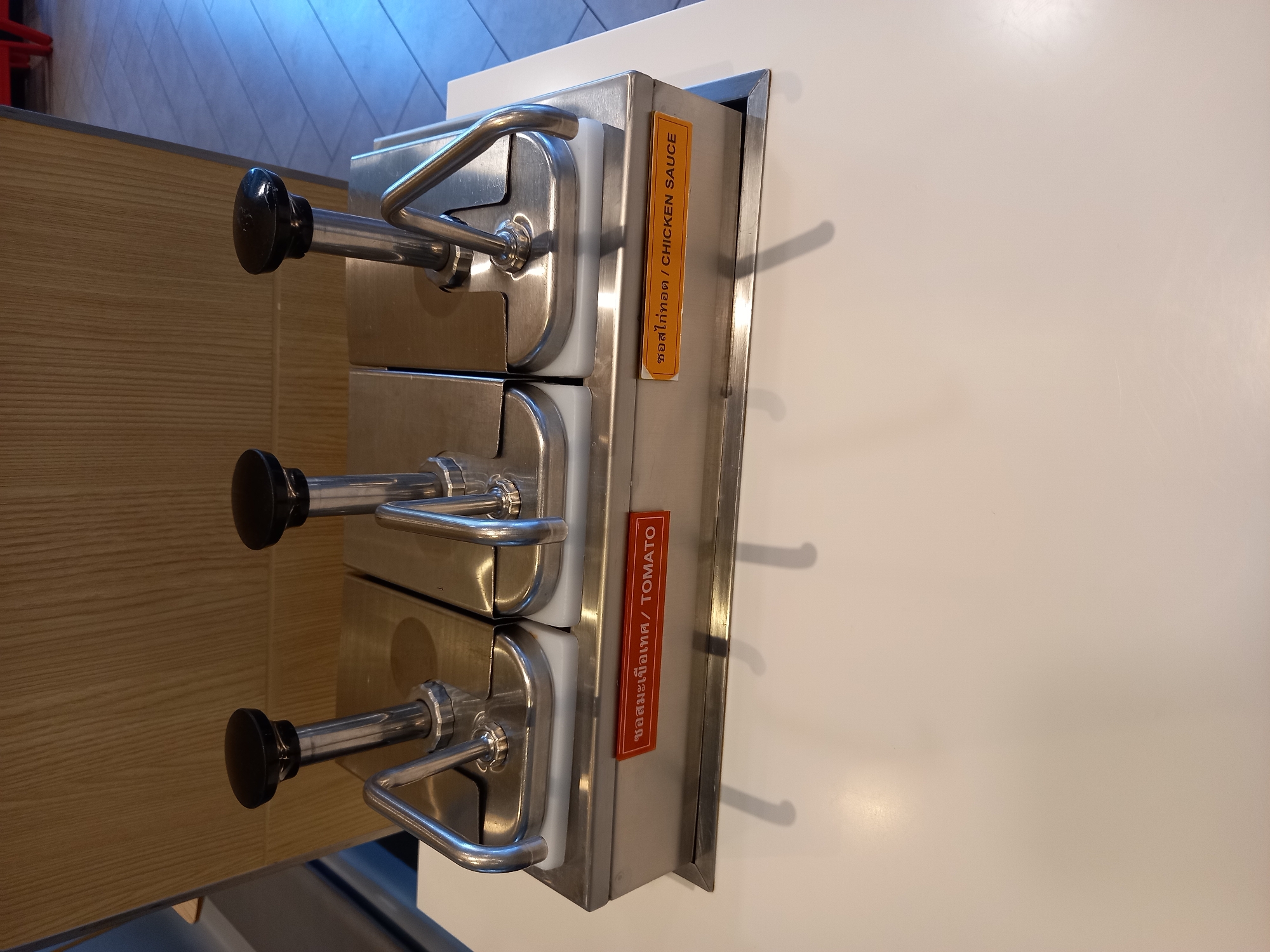 This media file shows a stainless steel condiment dispenser with three pumps. The dispenser is mounted on a white surface, with a wooden panel visible to the left. Each pump has a black handle. A vertical orange label is attached to the side of the dispenser, bearing the text "ซอสไก่ทอด / CHICKEN SAUCE". Below this, a vertical red label displays the text "ซอสมะเขือเทศ / TOMATO". The scene appears to be indoors, possibly in a restaurant or food service establishment. The lighting suggests it is daytime. There are no people visible, and the focus is on the dispenser and its labels. The overall impression is one of a functional food service appliance.