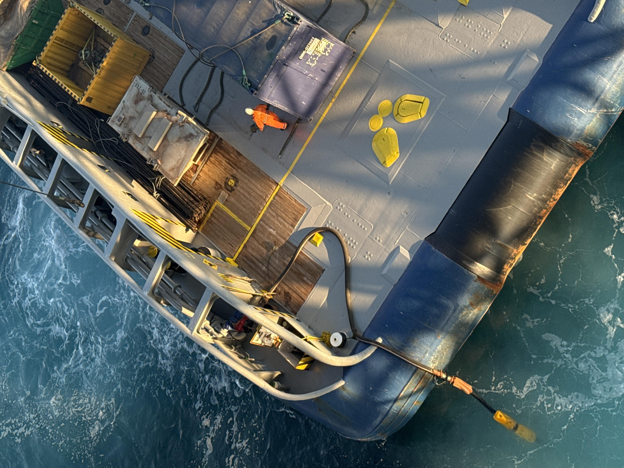The image shows an aerial view of the deck of a vessel at sea. A person in an orange jumpsuit is walking on the deck. There are several cargo containers on the deck, as well as some equipment and supplies. A hose extends from the vessel into the water, with a yellow marker visible on its submerged end. The water is a bright turquoise color and appears to be choppy, with white foam on the surface. The vessel has a blue hull and a grey deck with yellow markings. The scene is illuminated by sunlight, casting shadows across the deck. The location is not within a city.