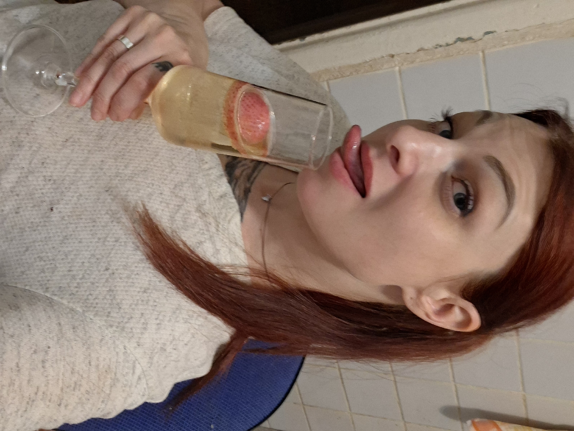 An adult female, fair-skinned with reddish-brown hair styled in a ponytail to the viewer's right, is depicted in an indoor setting. Her left eye, with a visible light blue/grey iris and mascara, looks forward. Her mouth is open, with a glossy, light red lip product, and her tongue is extended horizontally, slightly curled towards a clear, stemmed champagne flute. She holds the flute in her left hand, which bears a silver-colored band ring on the ring finger. The flute contains a translucent, yellowish-gold liquid with multiple thin, red strawberry slices floating within it. She wears a light-colored, possibly off-white or light grey, long-sleeved top featuring a knitted or textured pattern. A thin, silver-colored necklace with a small pendant is visible around her neck. A dark tattoo is partially visible on her left upper arm. The background includes light-colored square wall tiles with visible grout lines. To the left, a wall section exhibits peeling paint or plaster. On the far right, a portion of a dark blue, textured object is visible. A corner of a light-colored, possibly striped, fabric is present in the upper right. This scene is located in the city of Забалоцкі сельскі Савет, Belarus.