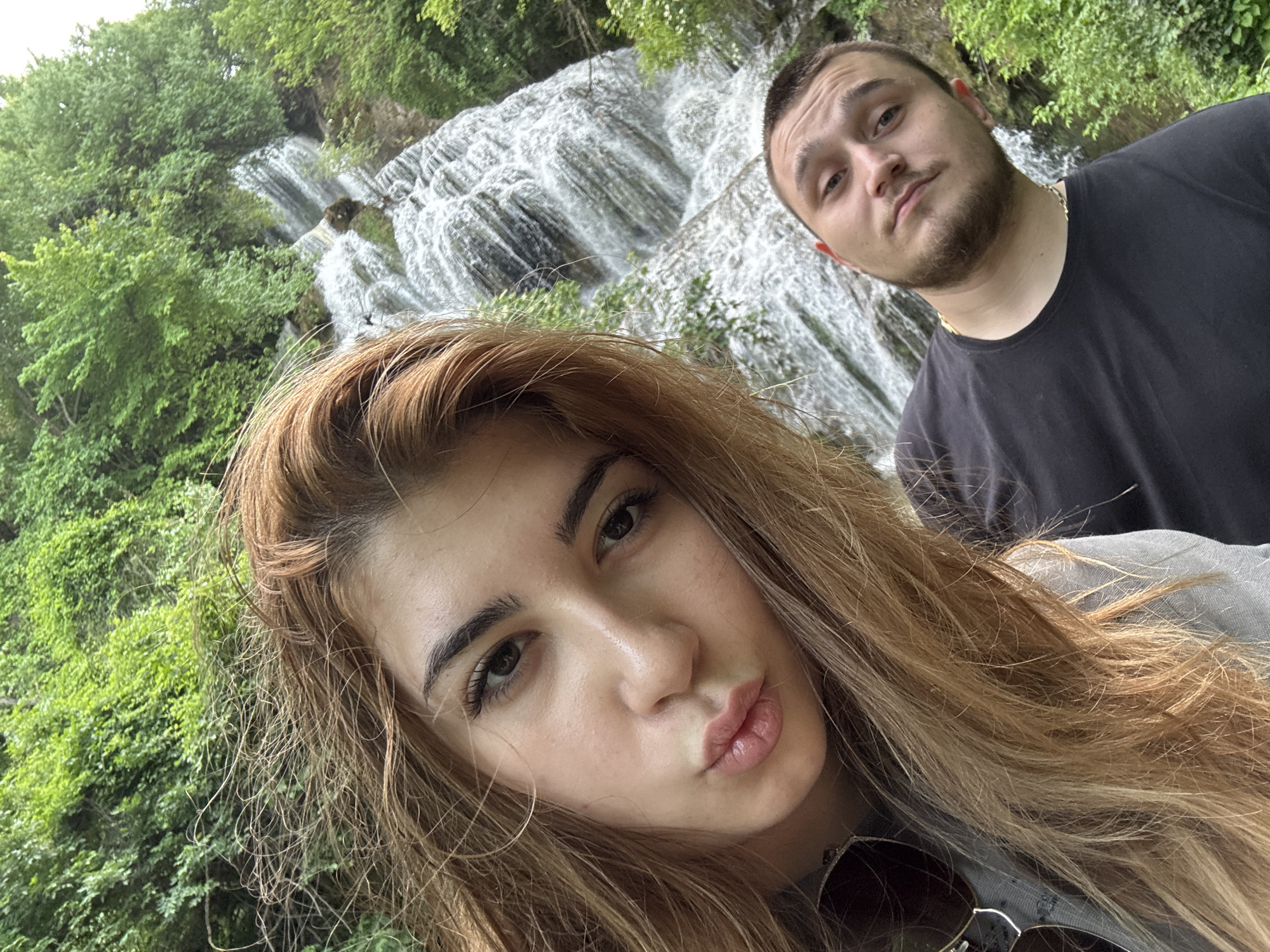 A selfie featuring a woman and a man is taken in Krushuna, Bulgaria, with a waterfall in the background. The woman is in the foreground with pouty lips and light brown hair. The man stands slightly behind her, looking into the camera. Both are likely tourists visiting the local natural attraction. The environment is lush and green, indicating a natural park or similar setting.