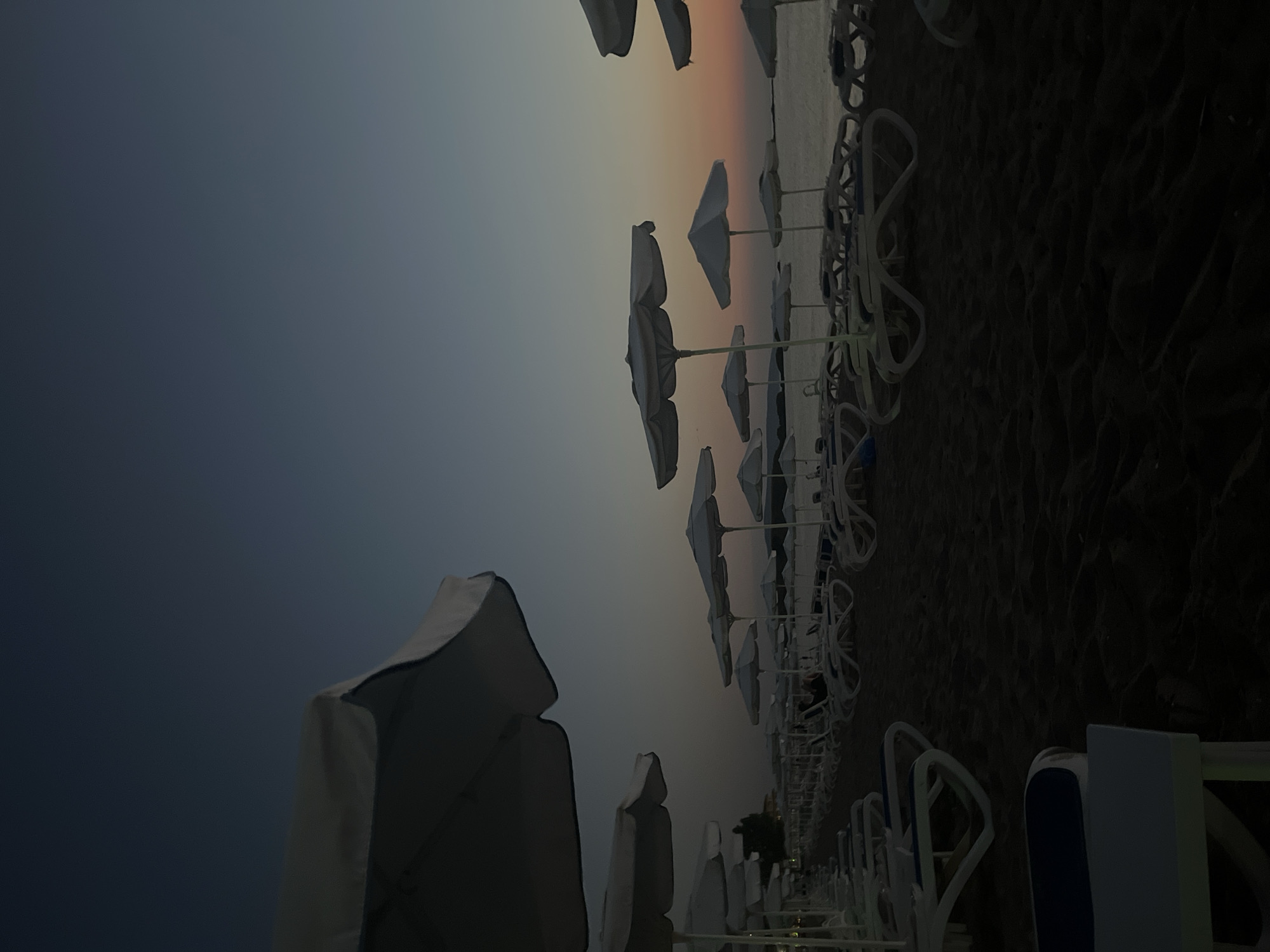 The image depicts a beach scene at dusk in Lozenets, Bulgaria. Rows of empty white lounge chairs and matching umbrellas are neatly arranged on the sand. The sky displays a gradient of deep blue at the top, transitioning to lighter shades of blue and then to warm orange and pink hues near the horizon, indicating sunset. The sea is visible in the background, appearing calm. The overall atmosphere is tranquil and serene, with the beach appearing deserted.