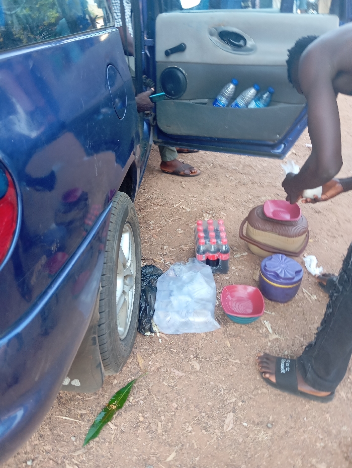The scene, located in Kadarko, Nigeria, depicts an outdoor environment on reddish-brown dirt ground next to a parked blue vehicle. The vehicle, a four-door model, has its passenger-side rear door open, revealing a light-colored interior door panel with a black speaker and three plastic water bottles stored in the door pocket.
Two individuals are present. One individual, partially obscured by the open door, is visible from the lower legs, wearing brown sandals, and appears to be inside the vehicle. The second individual stands outside the vehicle on the right side of the frame, clad in dark pants and black slides. This individual is bent over, holding and manipulating white plastic bags or items with both hands.
Various items are arranged on the ground next to the vehicle. These include a red carton holding multiple bottles, a black plastic bag, a clear plastic bag containing numerous white items, a stack of colored plastic bowls (red uppermost), a brown woven basket with a red lid, and a purple lidded container. A single green leaf rests on the dirt ground near the vehicle's rear wheel.