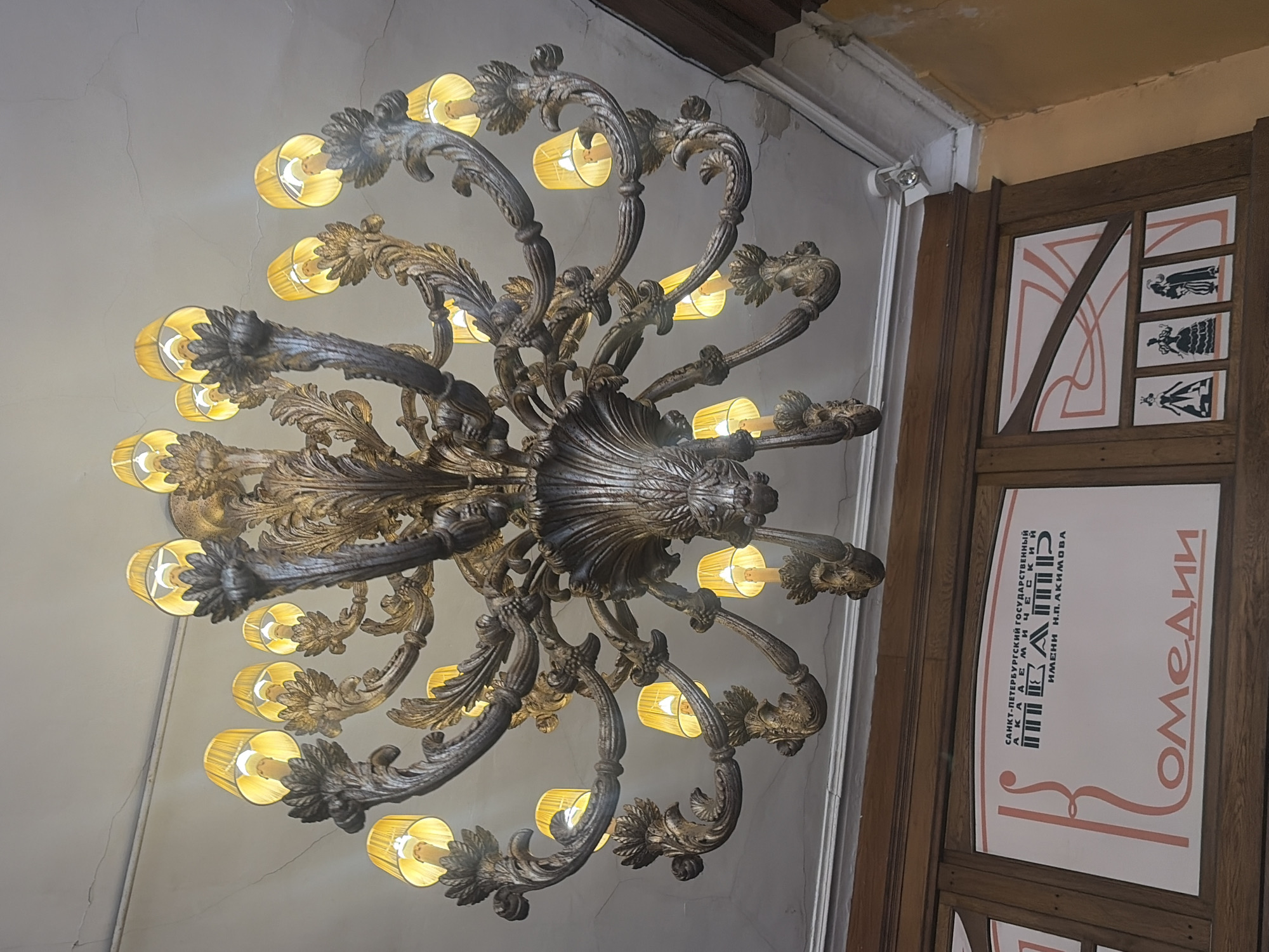 A grand, ornate chandelier with numerous golden arms and yellow-shaded lights hangs from a cracked white ceiling in a dimly lit interior. Below the chandelier, a wooden panel features a sign with Russian text and graphic elements. The sign reads "САНКТ-ПЕТЕРБУРГСКИЙ ГОСУДАРСТВЕННЫЙ АКАДЕМИЧЕСКИЙ ТЕАТР ИМЕНИ Н.П. АКИМОВА" and "Комедии". To the right of this sign, another panel displays several black silhouette figures in historical attire. The overall scene suggests an interior of a theatre or cultural venue.