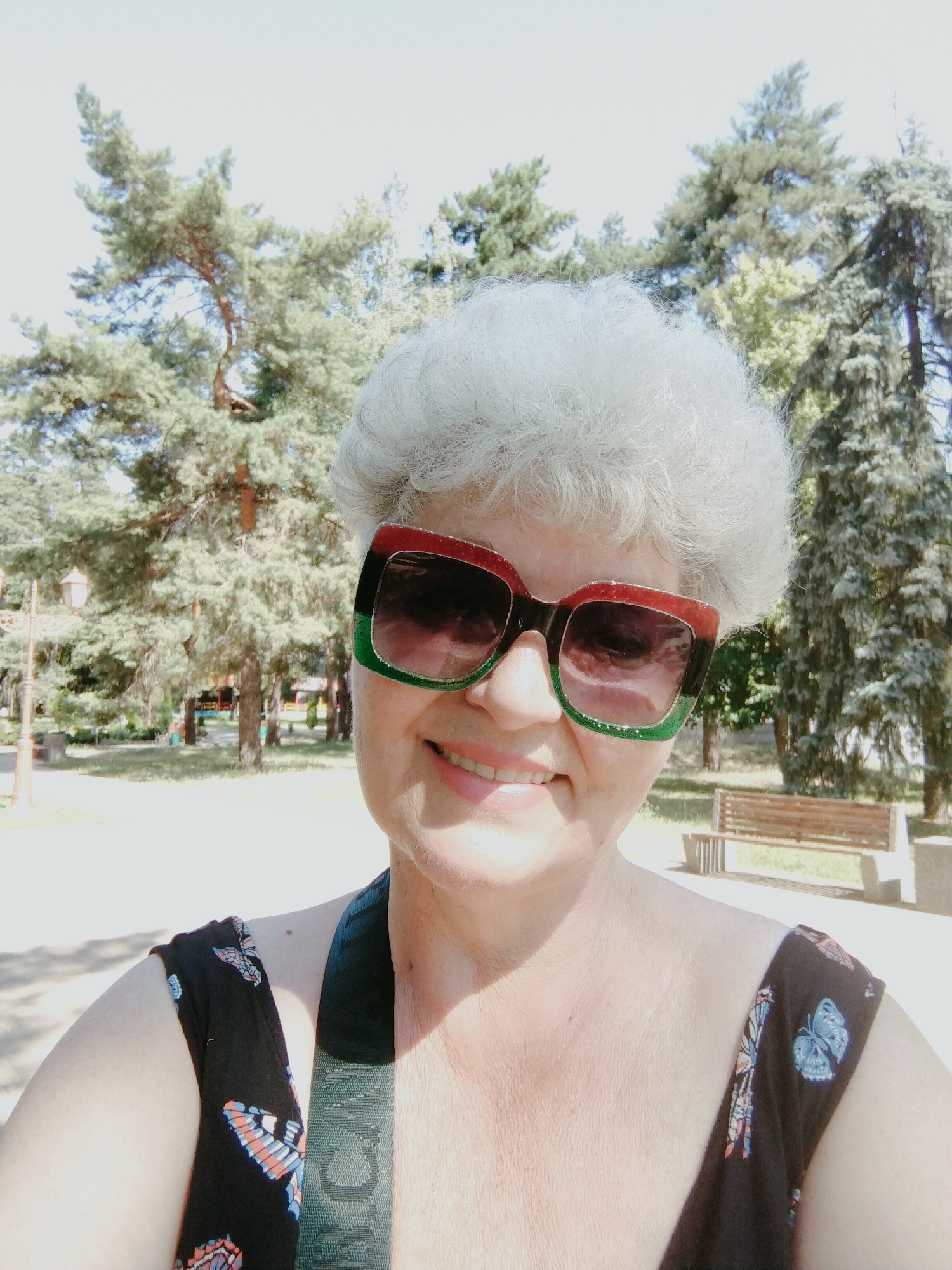 This is a medium shot, outdoor selfie of an older woman with short, white, curly hair. She is smiling and wearing oversized sunglasses with a red and green rim and dark lenses. She is wearing a black dress with a butterfly print and a dark strap over her right shoulder. The background shows a park with tall pine trees, a lamppost, a wooden bench, and sunlight filtering through the trees. The location is identified as Horishni Plavni, Ukraine.