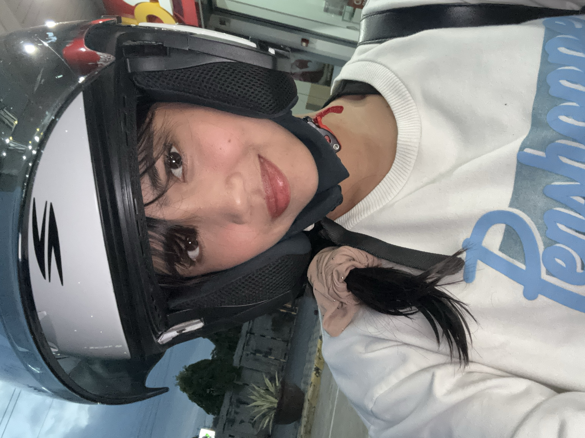 A young woman wearing a white sweatshirt and a motorcycle helmet is posing for a selfie. She has dark hair pulled back with a scrunchie and her hair is falling out of the ponytail. She is wearing light makeup and has a slight smile. The background appears to be outdoors, possibly at dusk, with a cloudy sky and some trees visible. There are buildings and a street in the background. The image is taken from a close-up perspective, focusing on the woman's face and upper body. There is no indication of the location being in Davao City, Philippines.