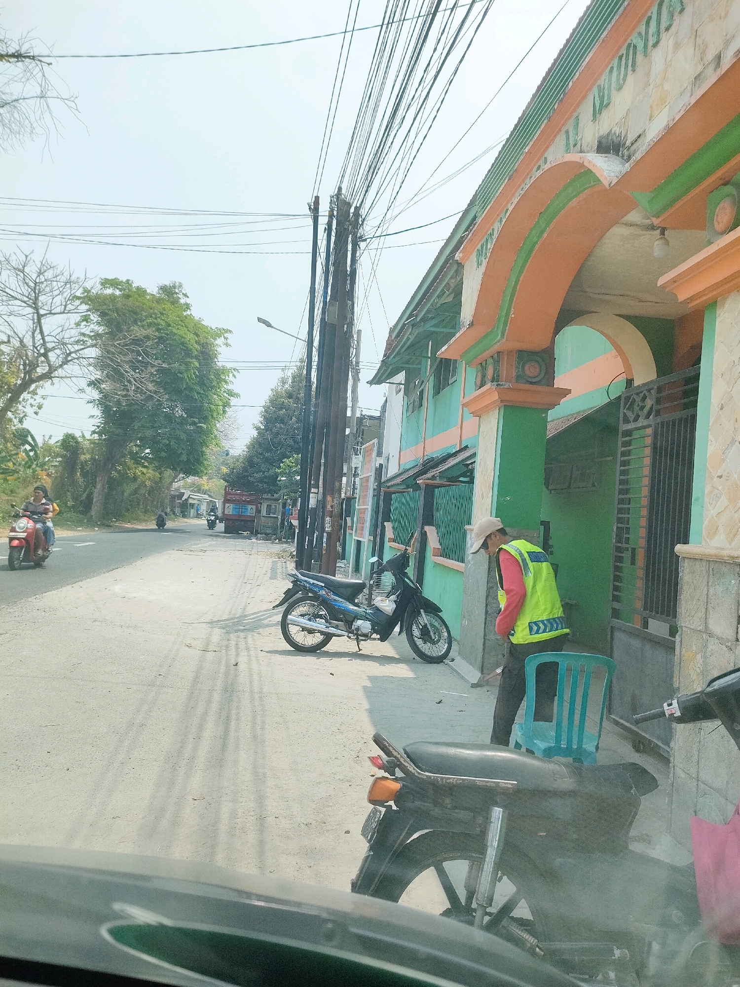 The image depicts a street scene in Pangkahwetan, Indonesia. On the right side of the frame, a building with an orange and green arched entryway is visible. A person wearing a bright yellow vest and a cap is standing near the entrance. Two motorcycles are parked on the side of the road; one is a blue motorcycle to the left of the person in the yellow vest, and another is a dark-colored motorcycle in the foreground. In the background, a red truck and other vehicles are on the road, and trees line the street. Overhead, numerous electrical wires are strung between poles. The overall environment appears to be a moderately busy street on a bright, sunny day.