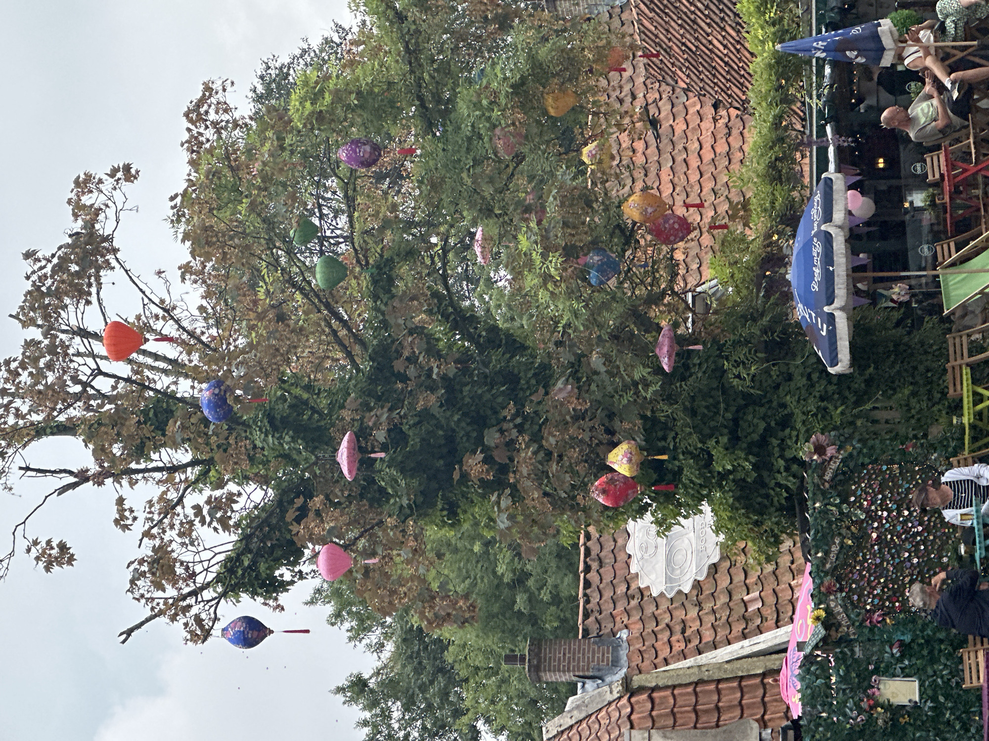 The image shows a tree adorned with numerous colorful lanterns and decorative umbrellas. Several people are seated at tables outdoors, some appearing to be dining or socializing. The background features red-tiled rooftops, characteristic of Amsterdam's architecture, and lush green foliage. The sky is overcast, suggesting a cloudy day. The overall scene depicts a charming outdoor seating area, possibly a cafe or restaurant, decorated for a festive occasion.