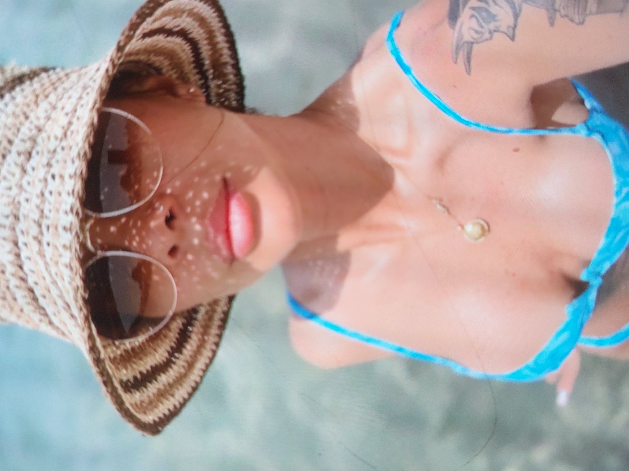 An overhead shot captures a person, likely a woman, relaxing in clear, light blue-green water. The image is rotated, with her head positioned towards the left of the frame, looking up towards the camera. She wears a wide-brimmed straw hat with alternating light and dark brown stripes, and round, dark-tinted sunglasses. Her lips are slightly parted.
Strong sunlight illuminates the scene, casting dappled patterns of light and shadow across her face and neck, possibly due to reflections from the water or light filtering through the hat's weave. She is wearing a light blue bikini top, and a delicate necklace with a small, round, light-colored pendant hangs on her chest. A black and grey tattoo, depicting what appears to be an animal or mythological creature's head, is visible on her left shoulder/upper arm. Her fingernails are painted white. The setting suggests a sunny day at a beach or pool in a location like Tsarevo, Bulgaria, emphasizing relaxation. No text is visible.