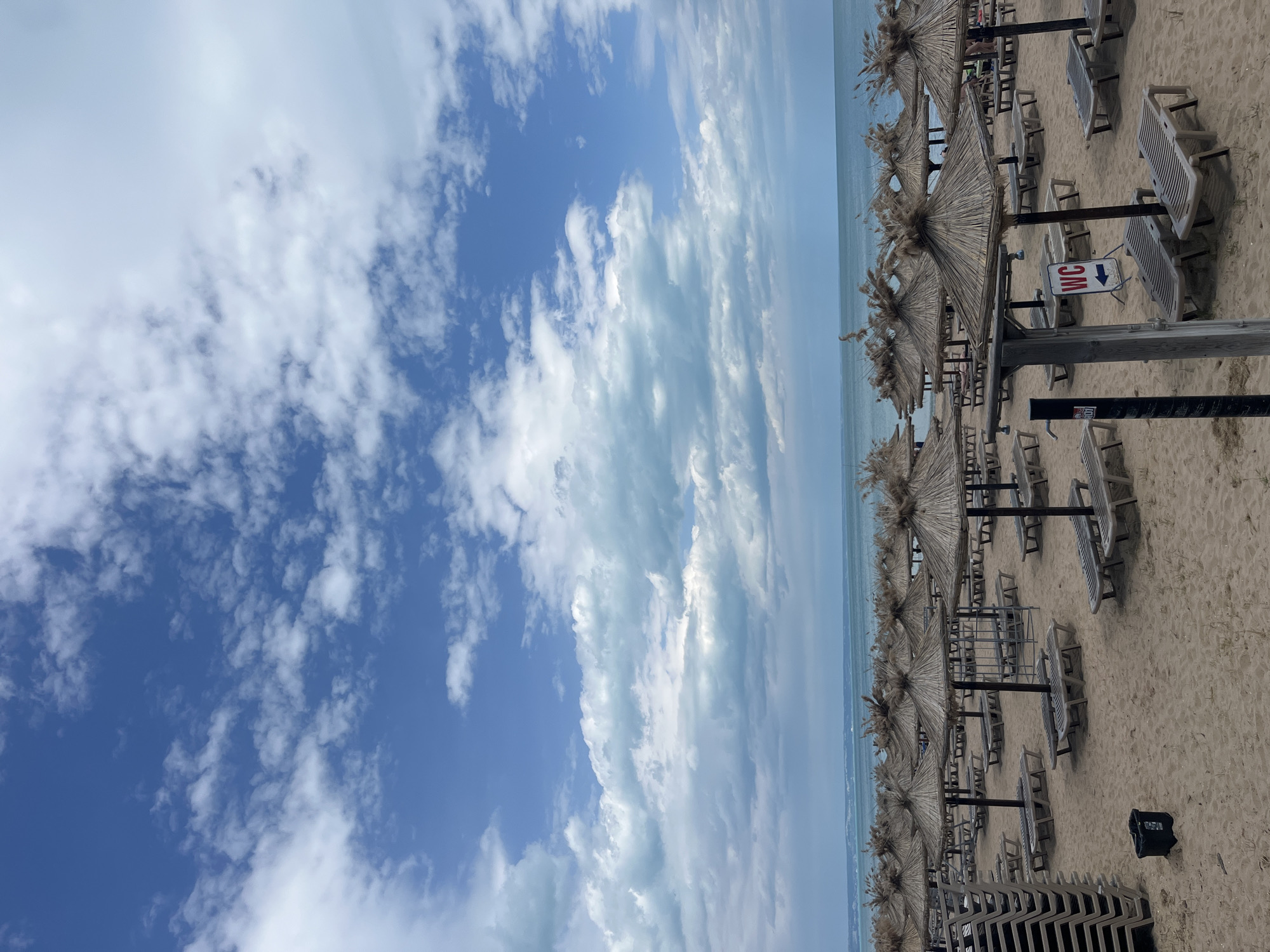 The image shows a beach in Varna, Bulgaria, viewed from a slightly elevated perspective, rotated 90 degrees counter-clockwise. The scene is dominated by rows of beach umbrellas and deck chairs, arranged in neat lines along the sandy beach. The umbrellas are made of natural materials, with thatched or straw coverings. A blue sea meets a blue sky covered in white clouds. A sign with "WC" and a downward-pointing arrow is seen. The scene is mostly devoid of people.