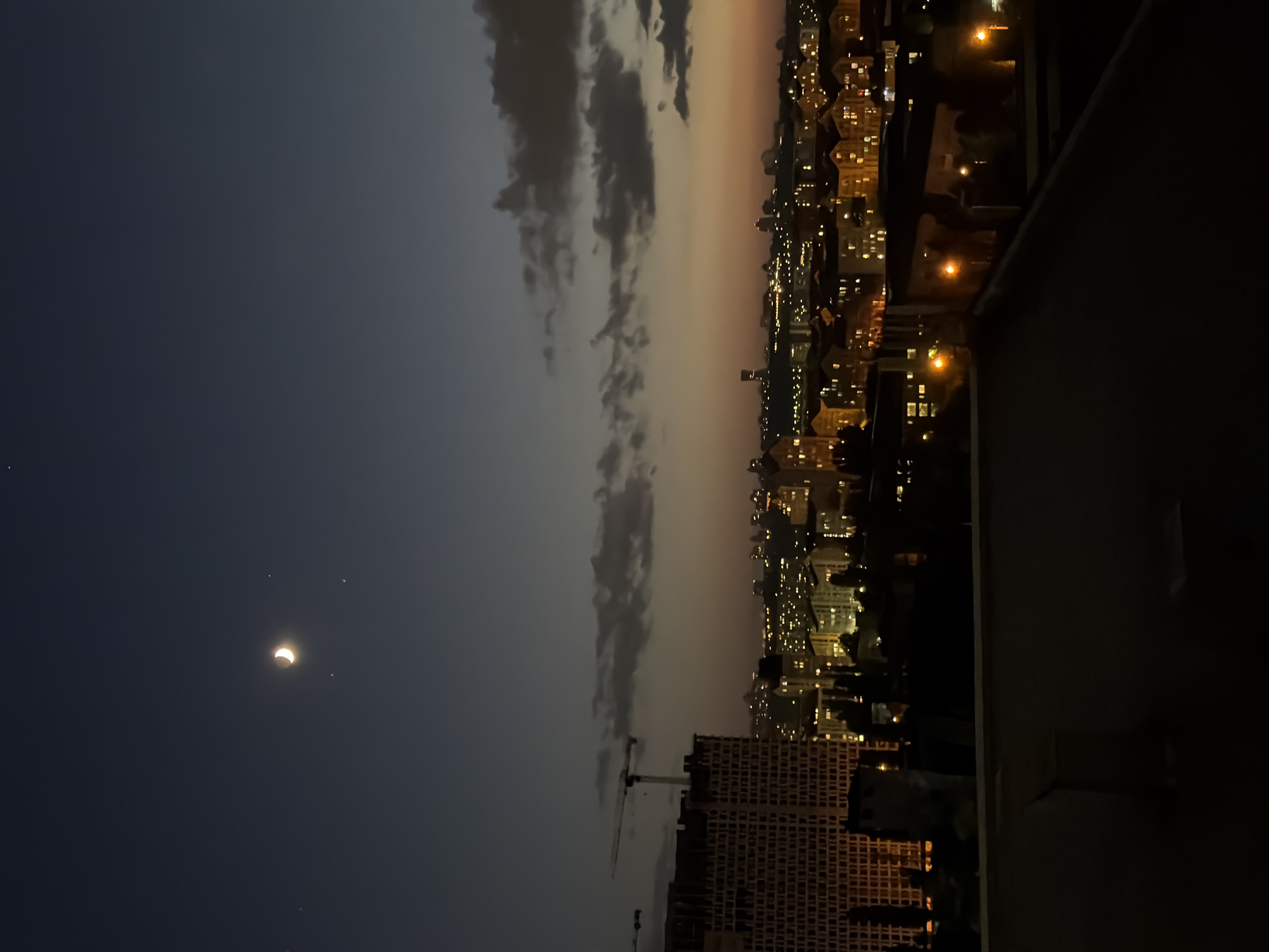 This media file captures a nighttime view of a city, likely Kyiv, Ukraine, from a high vantage point. The scene is dominated by the urban landscape, with numerous buildings illuminated by warm, yellow lights, suggesting a developed residential or commercial area. The image is oriented vertically, with the horizon running along the left side of the frame.
In the dark sky, a crescent moon is clearly visible, with two stars nearby. The moon's sliver of light is illuminated by the sun, indicating a phase of the lunar cycle. Dark clouds are scattered across the sky, partially obscuring the city lights in some areas and creating a dramatic contrast with the illuminated cityscape.
In the foreground, a dark, possibly building facade or balcony railing, takes up a significant portion of the right side, framing the view of the city. The overall atmosphere is tranquil and contemplative, hinting at the end of the day or early evening. The presence of artificial lights and construction cranes suggests an active, modern urban environment. No text is visible in the image.